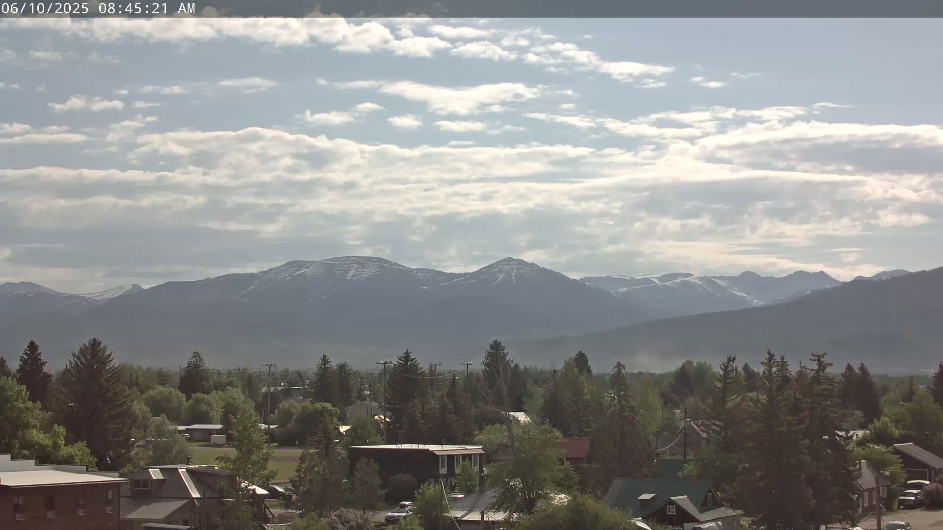A partly cloudy day reveals snow-capped mountains in the distance behind a town filled with evergreen trees and houses.