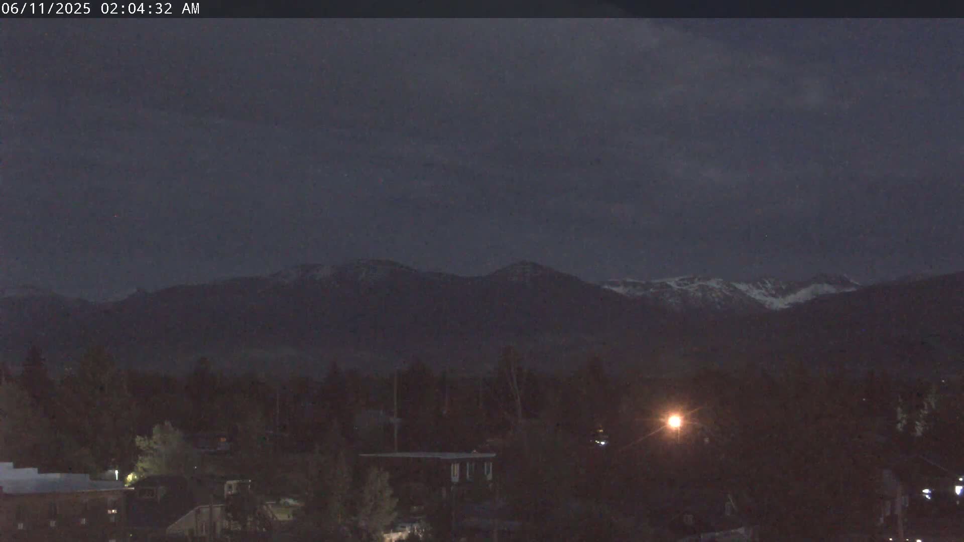 A nighttime view shows snow-capped mountains in the distance behind a dark, tree-filled landscape illuminated by a few streetlights under a dark, cloudy sky.