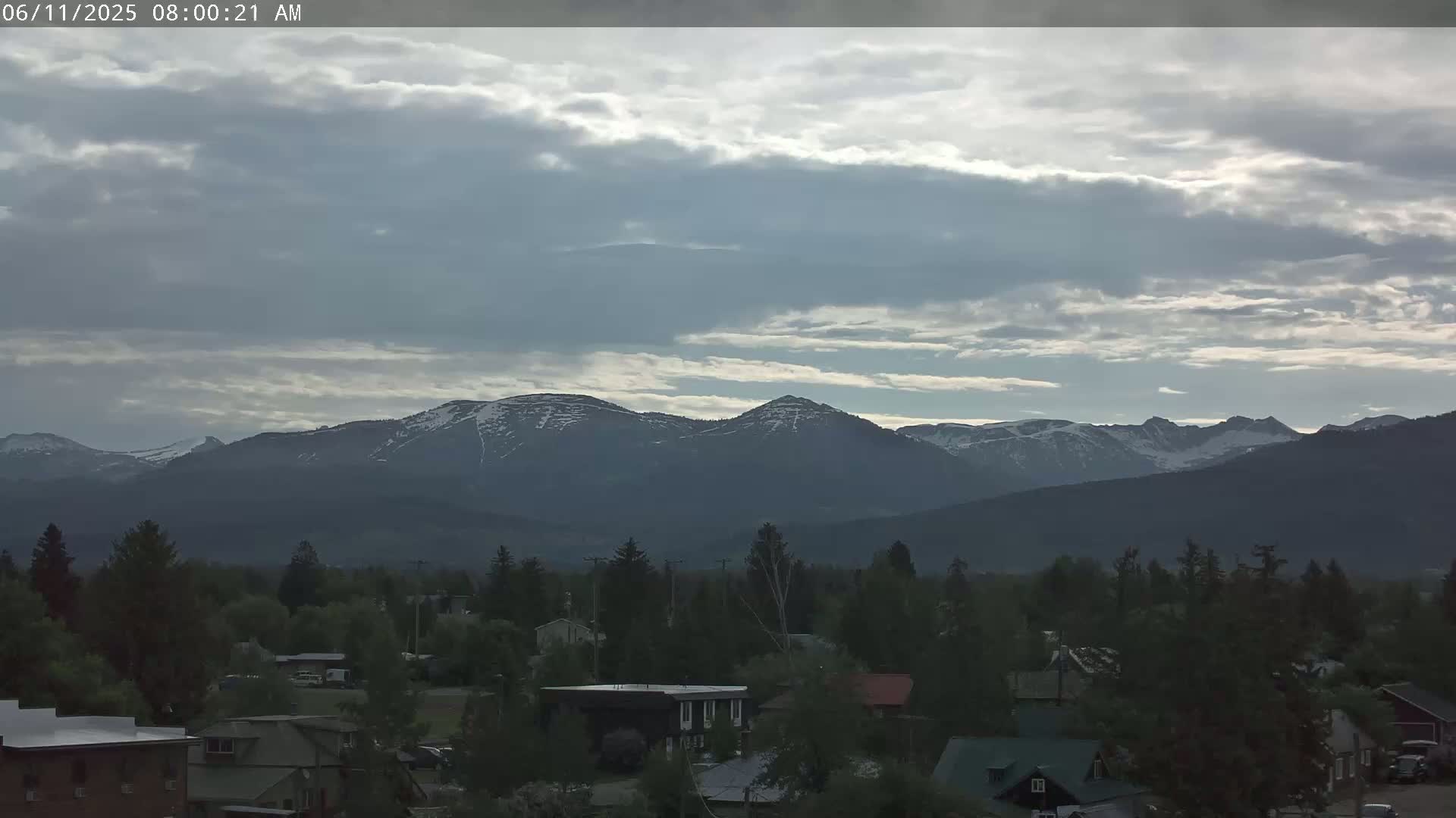 A partly cloudy sky overlooks a small town nestled among dark green trees and mountains with snow-capped peaks in the distance.