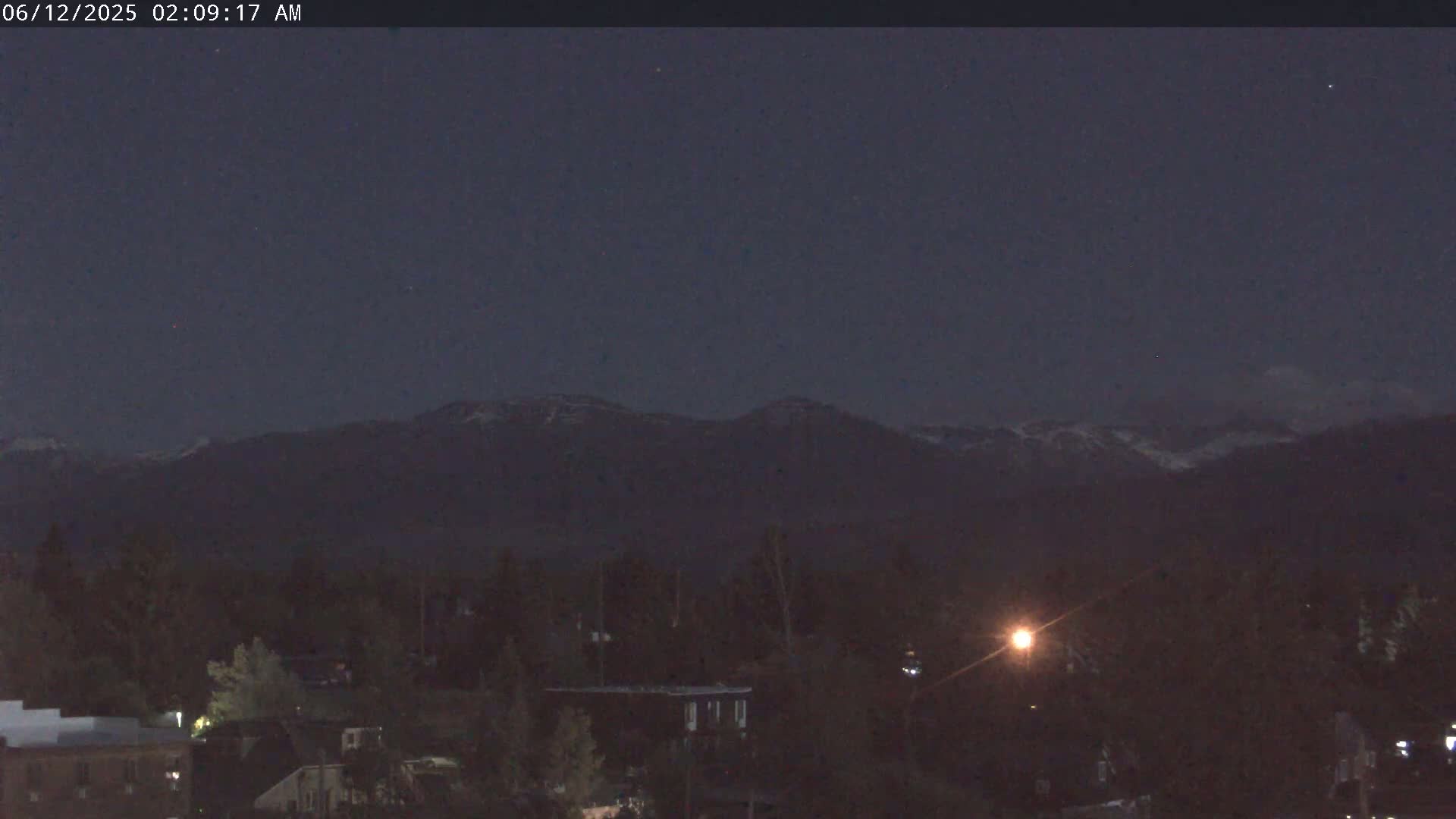 A nighttime view shows a snow-capped mountain range in the distance behind a dark, sparsely lit town under a clear, dark sky.