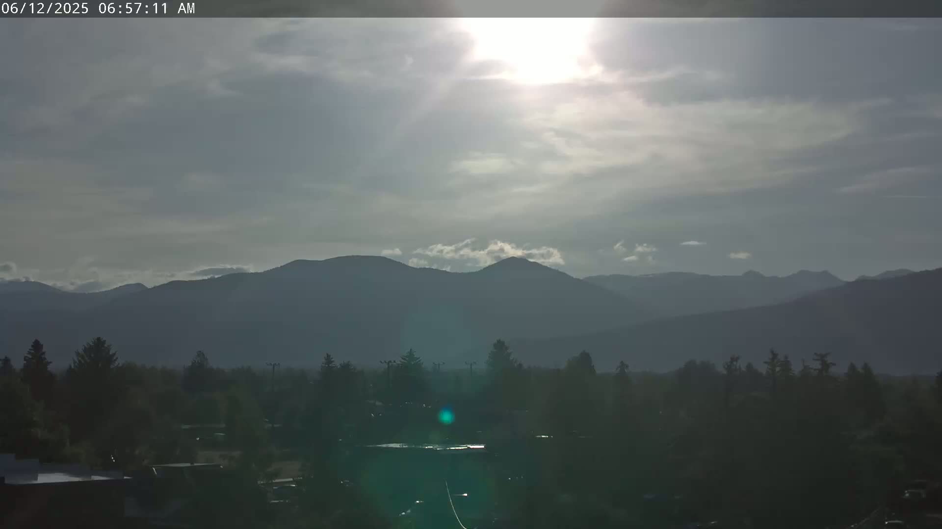 A partly cloudy morning sun shines over a line of dark, distant mountains and a foreground of dark, silhouetted trees and buildings.