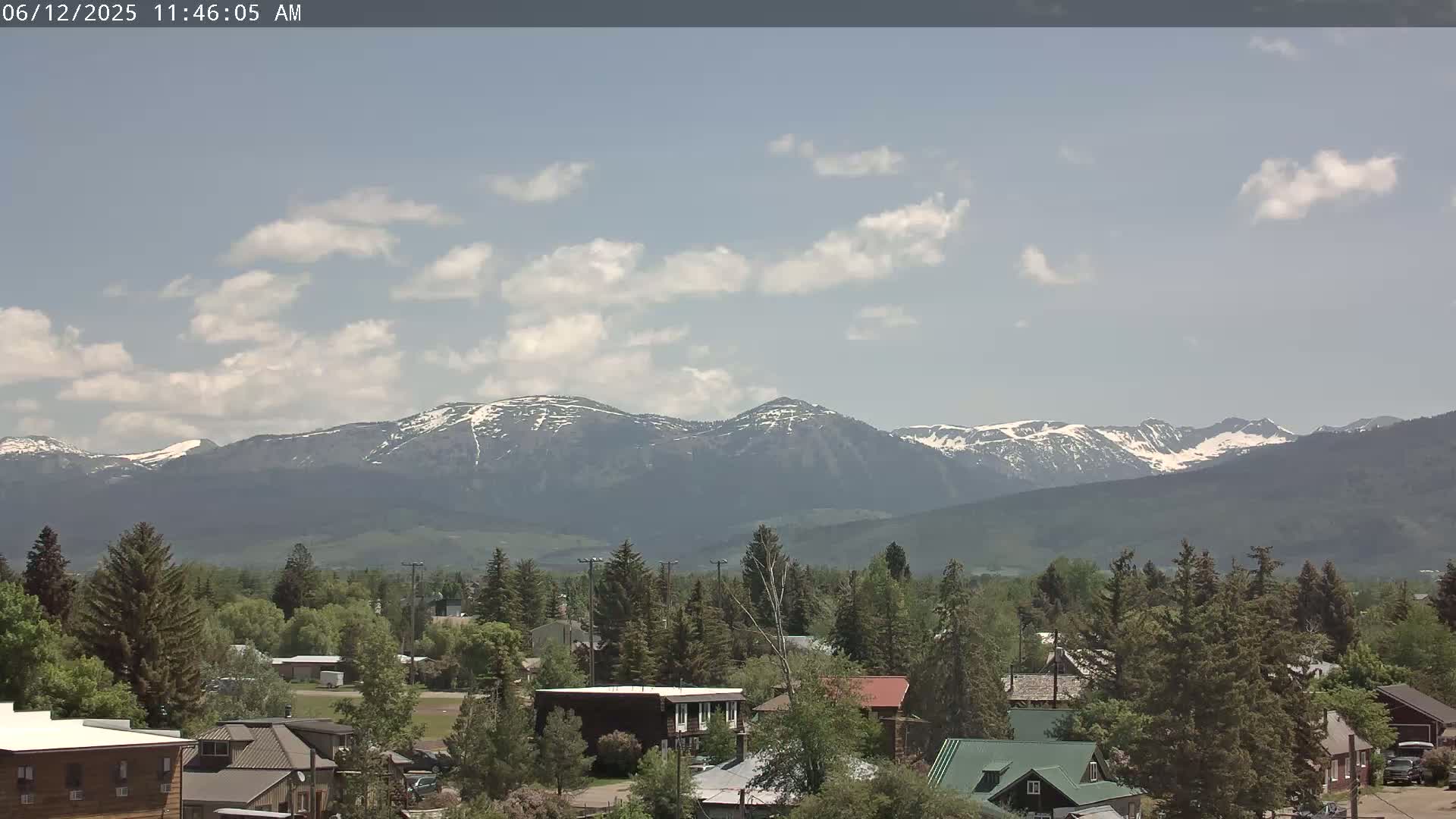 A partly cloudy day reveals a small town nestled amongst evergreen trees with snow-capped mountains in the background.