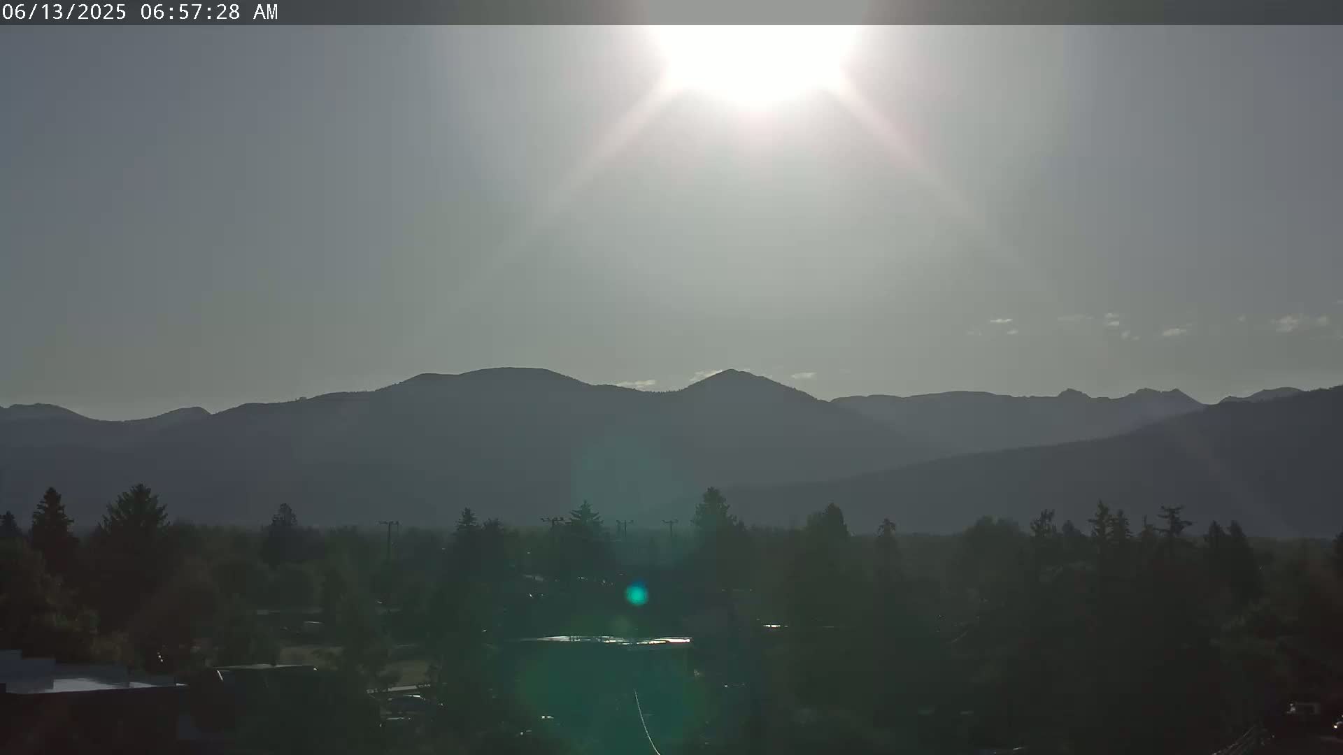 A bright sun shines over a hazy landscape of dark, distant mountains and a foreground of trees and buildings.