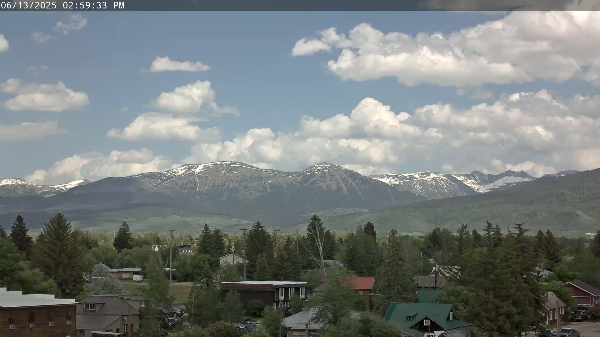 A partly cloudy day shows a small town nestled among green trees and hills, with snow-capped mountains in the distance.
