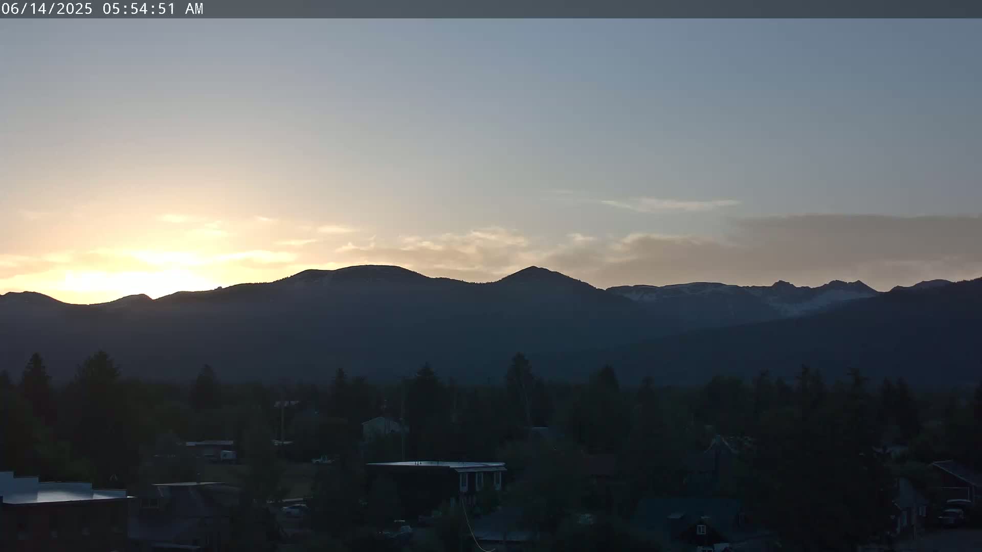 A sunrise illuminates snow-capped mountains behind a dark silhouette of a town under a mostly clear sky.