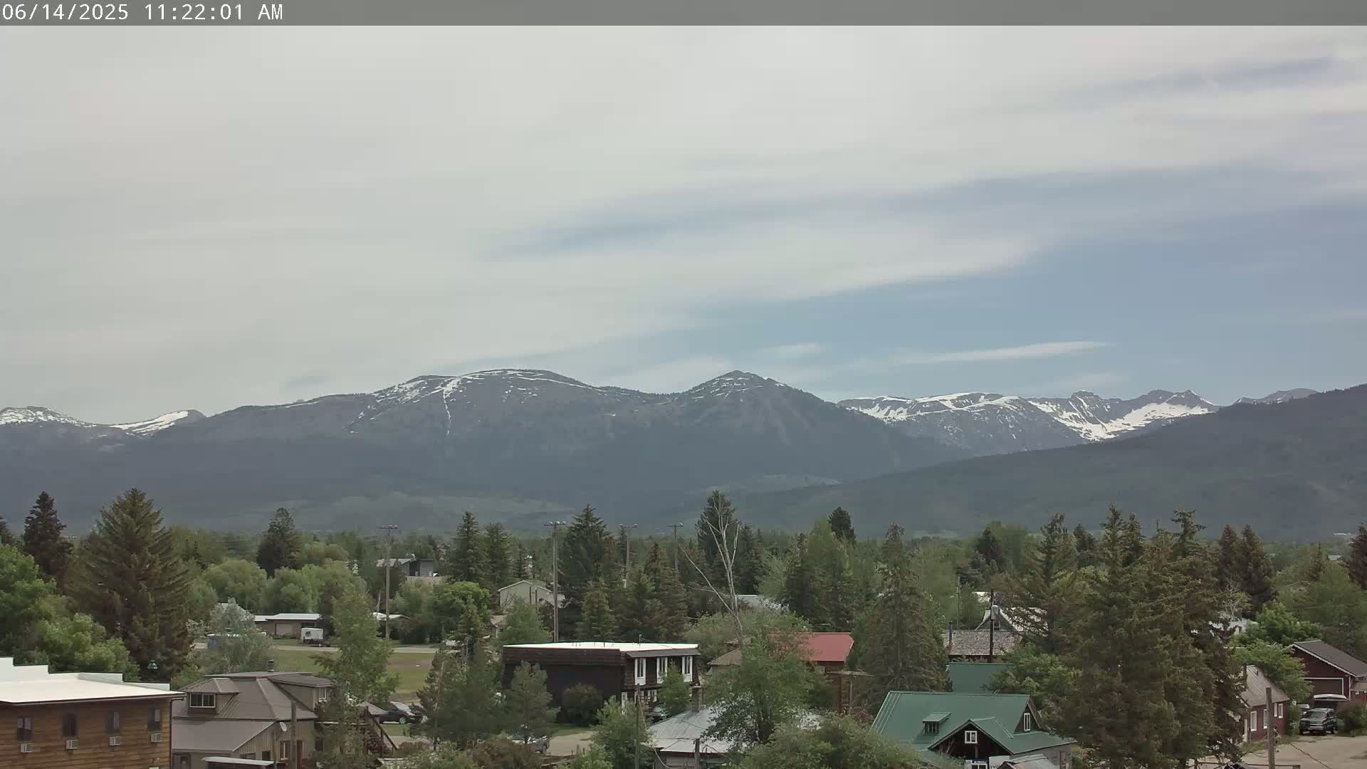 A small town nestled among green trees and shrubs, with snow-capped mountains visible in the distance under a partly cloudy sky.
