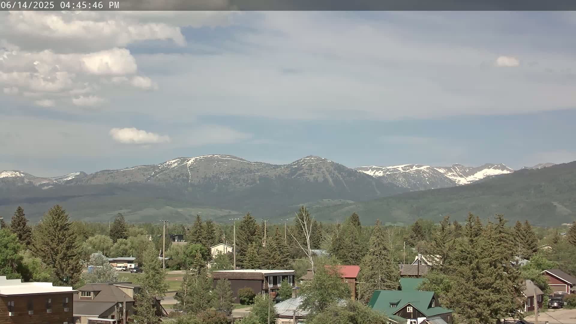 A small town nestled among green trees and shrubs, with snow-capped mountains visible in the distance under a partly cloudy sky.