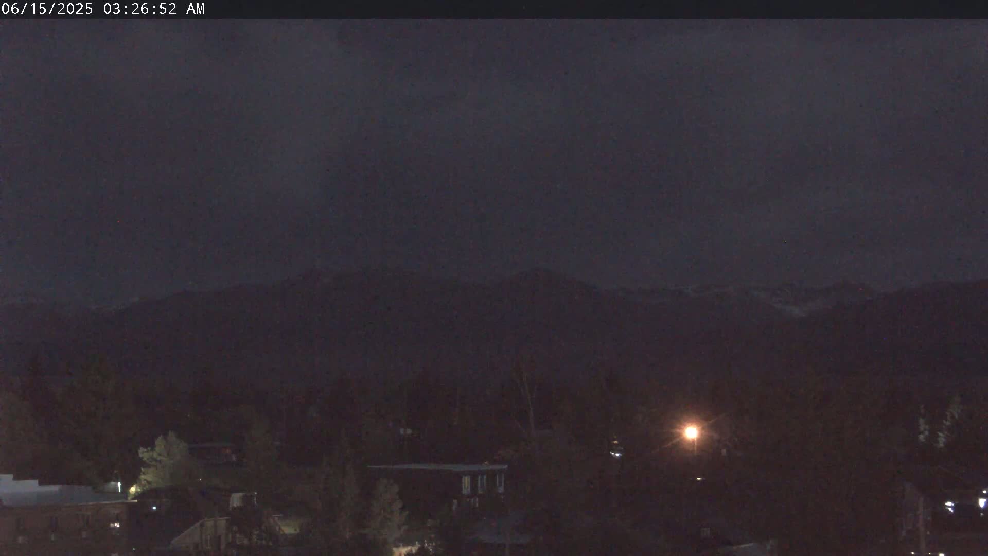 A nighttime view shows dark mountains in the distance, with a small town of dark buildings and trees in the foreground under a cloudy sky.
