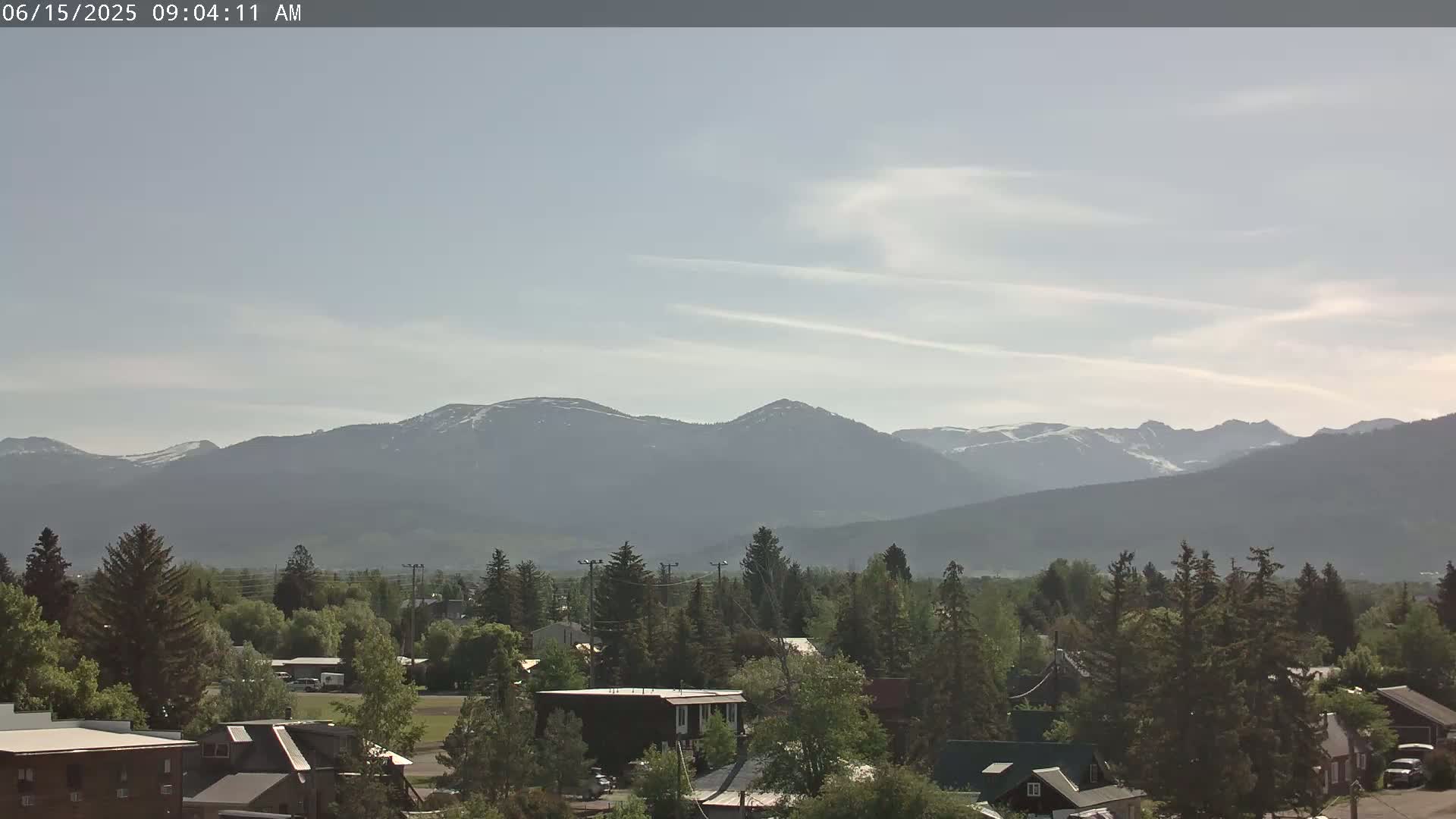 A small town nestled among evergreen trees, with a hazy mountain range showing patches of snow in the background under a mostly clear sky.