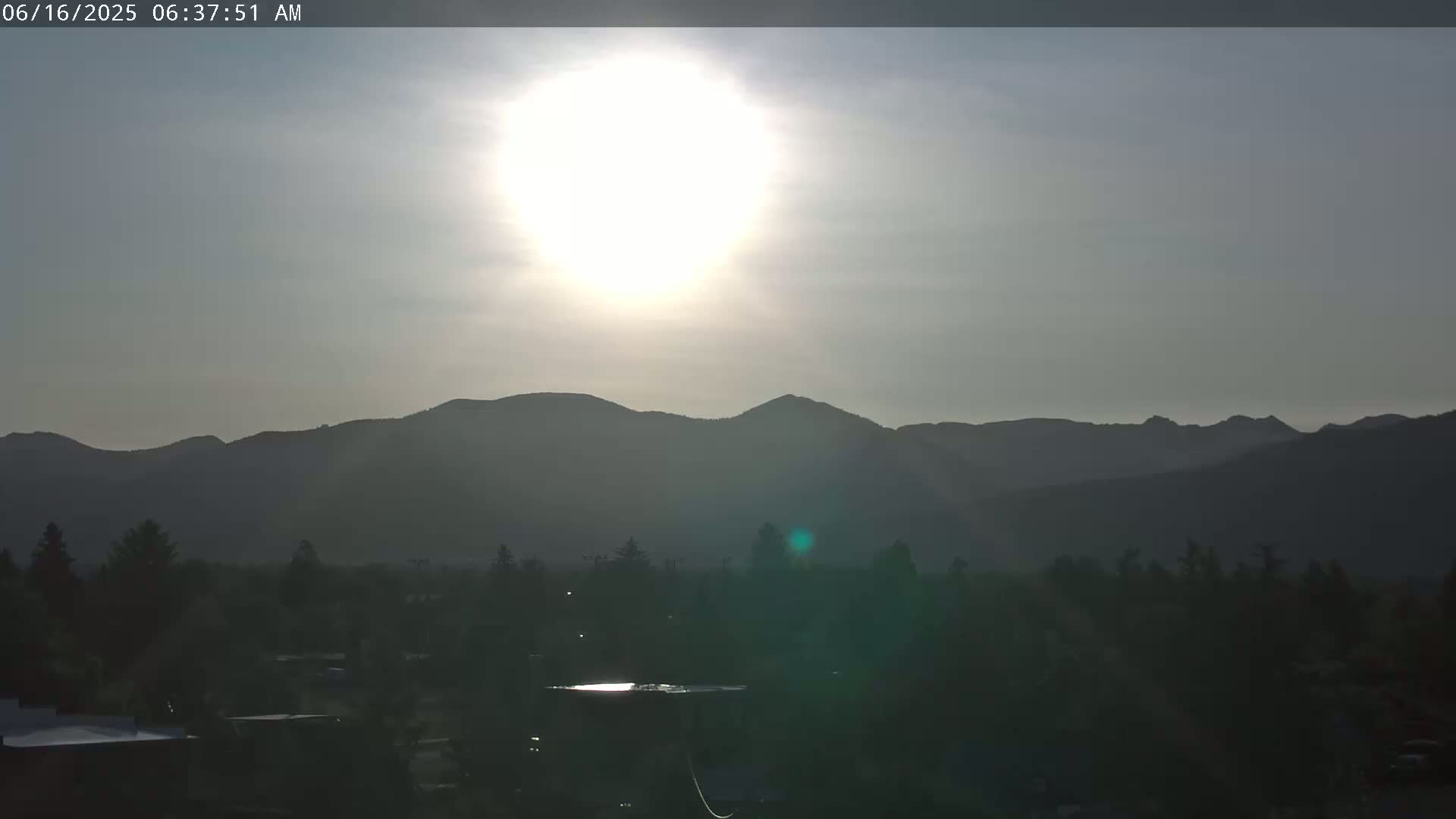 A bright sun shines in a hazy sky over a dark mountain range and a silhouetted town.
