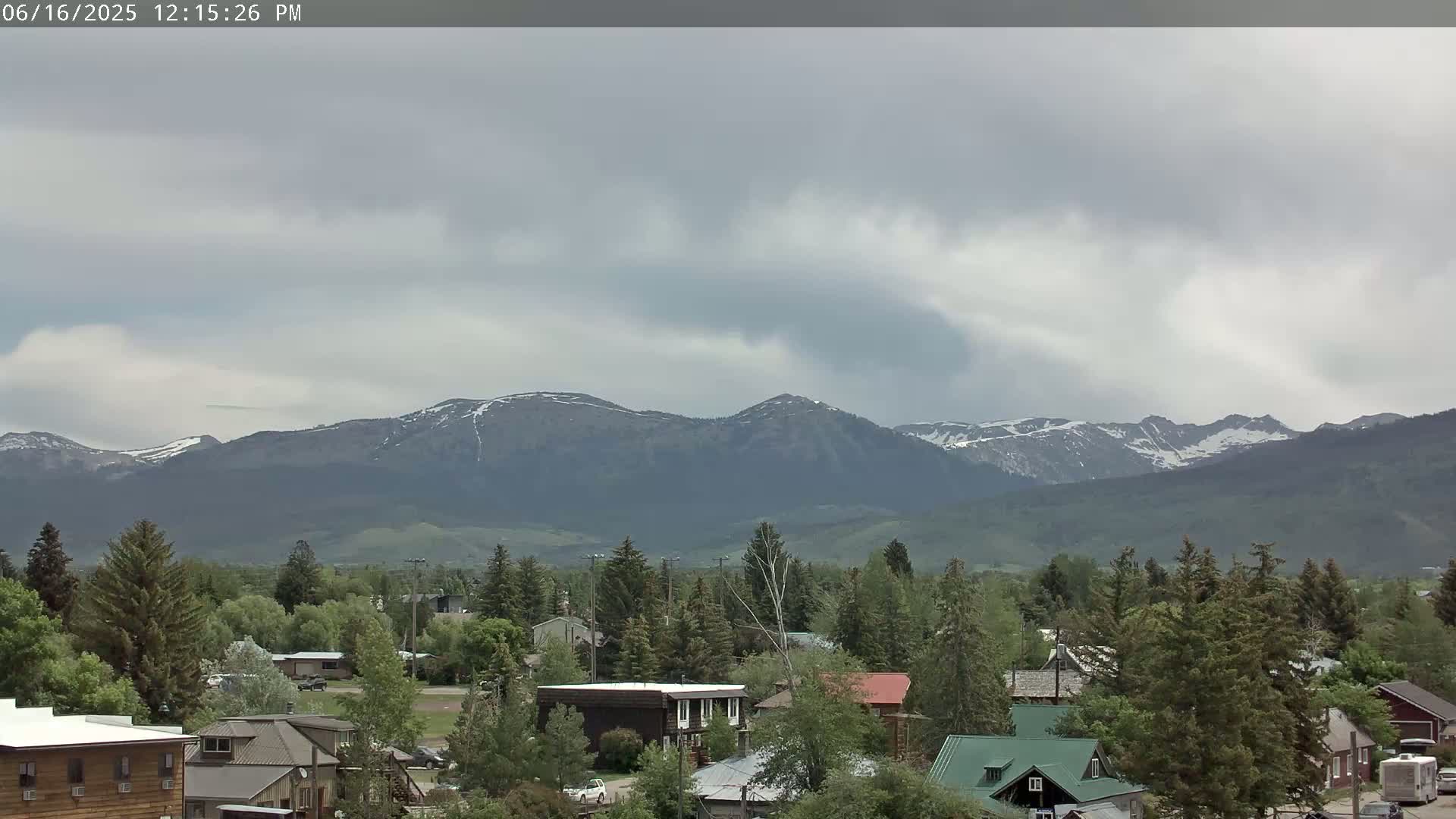 A small town nestled among green trees sits before a mountain range with snow-capped peaks under a cloudy sky.