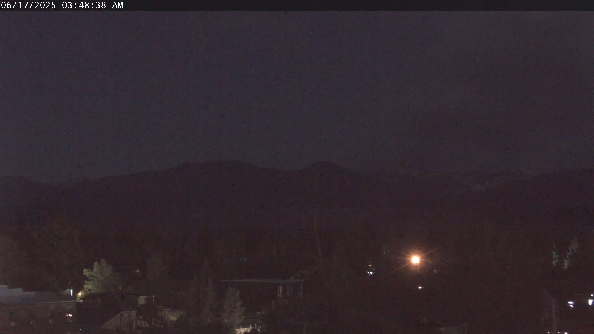 A nighttime view of dark mountains in the distance, with some sparsely lit buildings and trees in the foreground under a clear, dark sky.