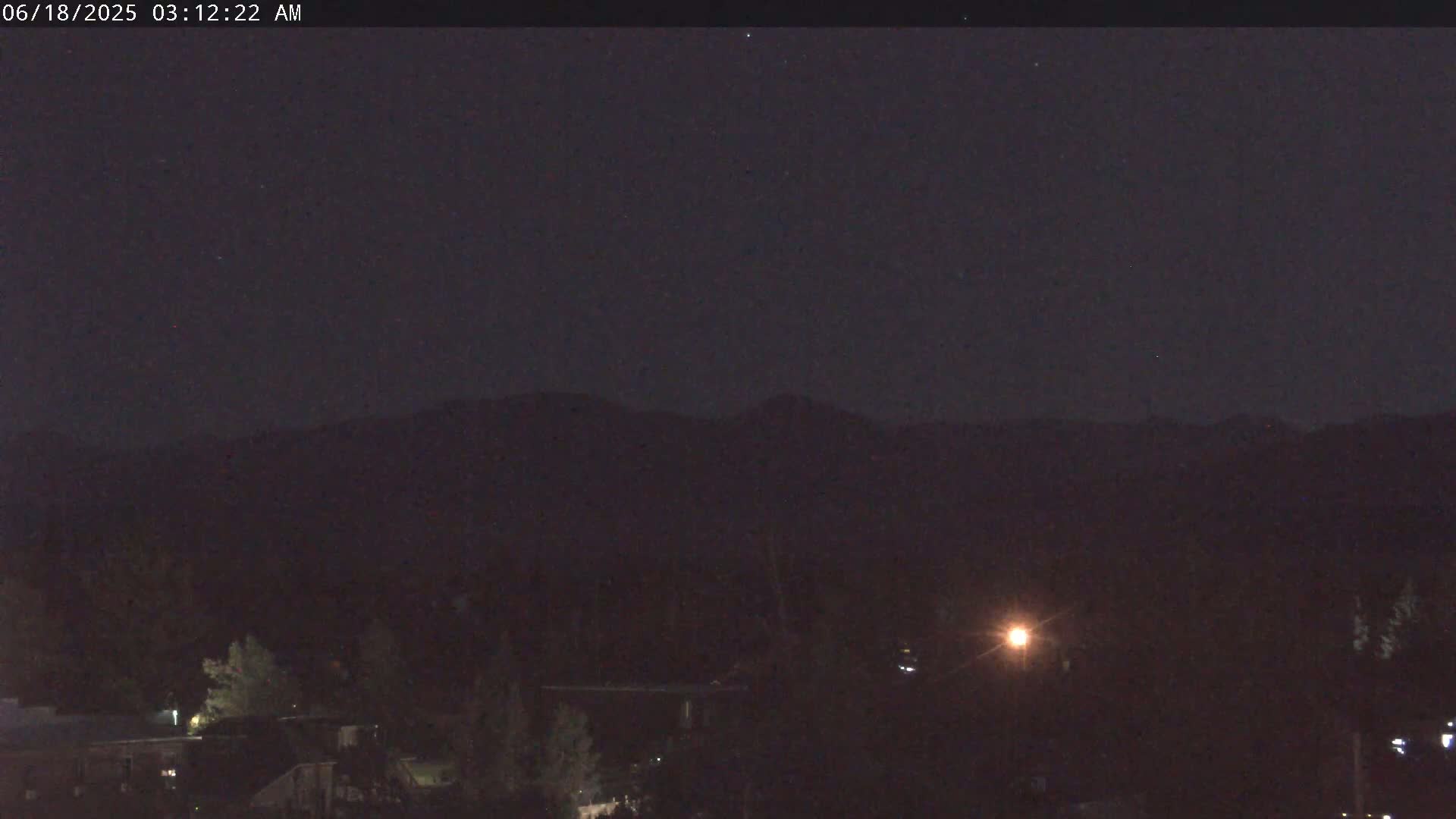 A dark, nighttime scene shows a distant mountain range silhouetted against a clear sky above a low-lying area with some trees and buildings.