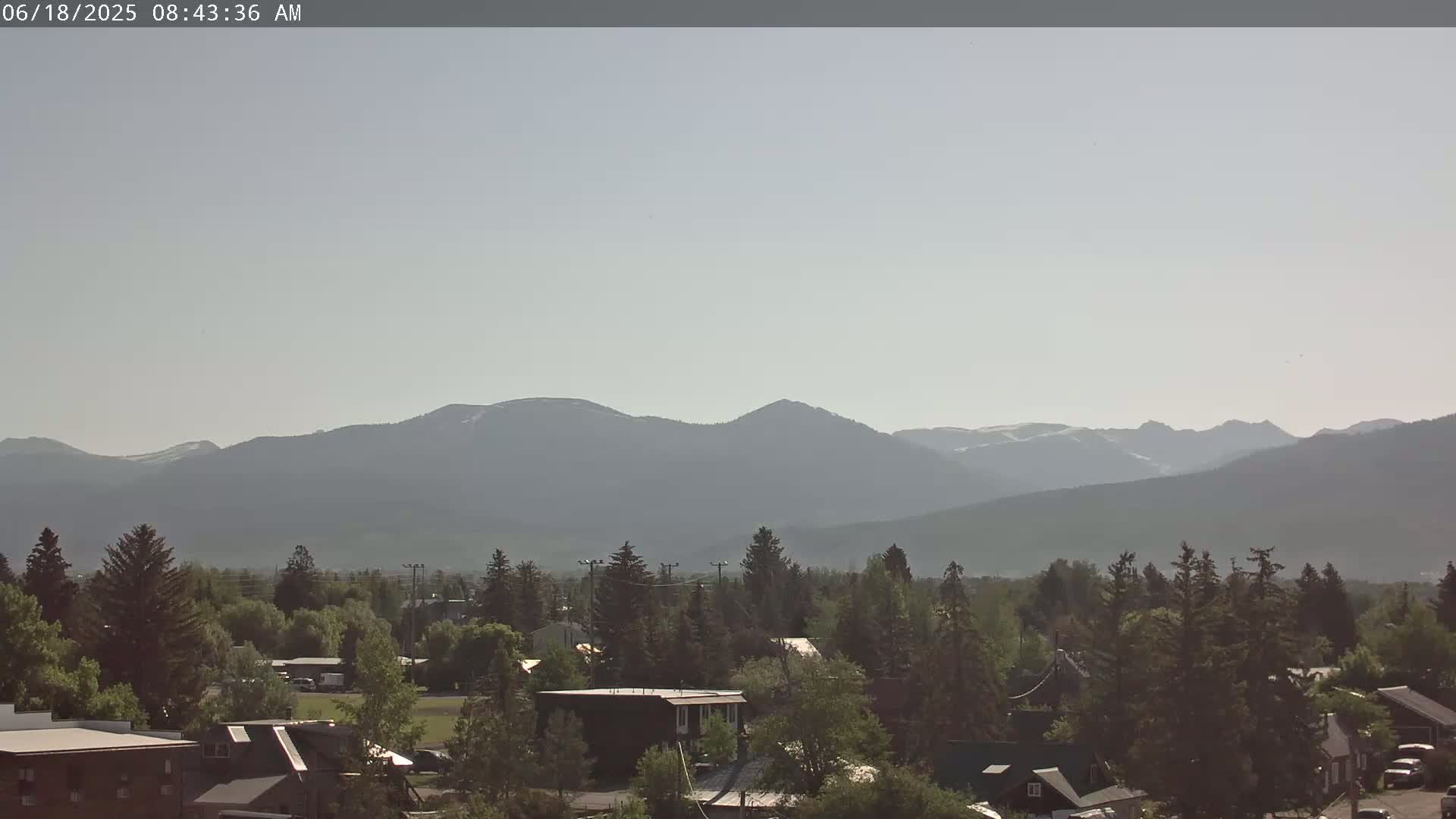 A hazy, sunny morning reveals a small town nestled among evergreen trees with a mountain range in the background, partially snow-capped.