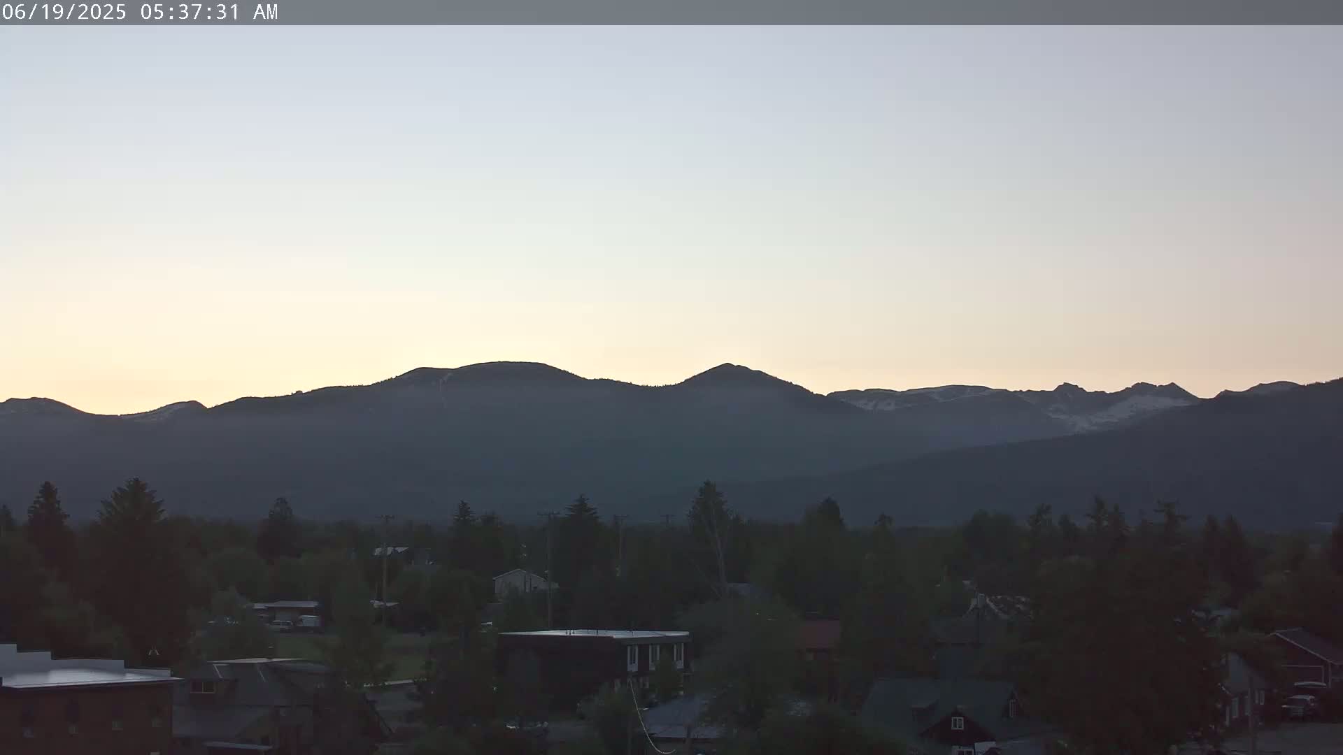 A pre-dawn, pale-orange sky illuminates a town nestled at the base of a dark, snow-dusted mountain range.