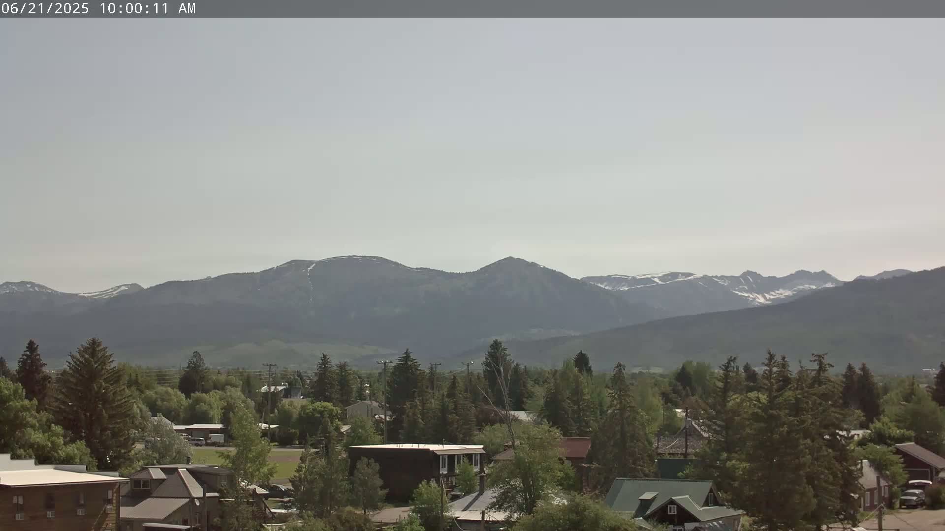 A hazy, sunny day reveals a small town nestled among evergreens, with snow-capped mountains visible in the distance.