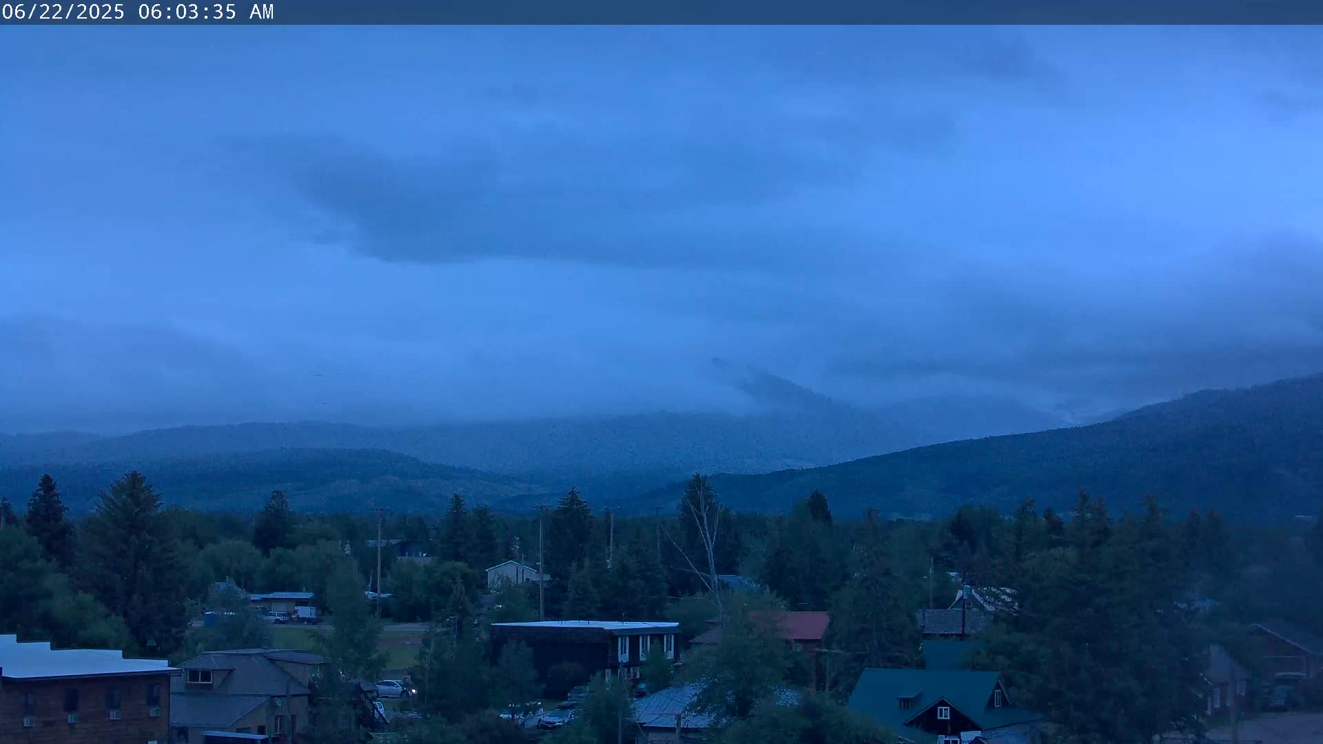 A small town nestled amongst trees at dawn, with hazy, dark blue skies and distant, cloud-covered mountains.