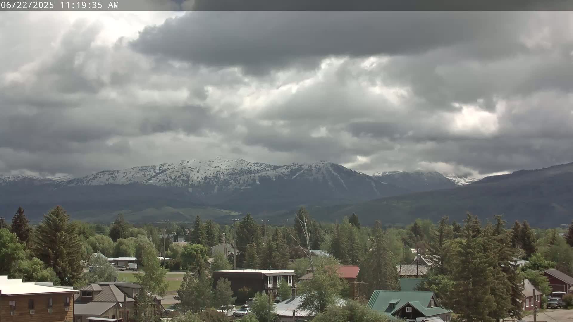 A small town with houses and evergreen trees sits in the foreground, with snow-capped mountains in the background under a cloudy sky.
