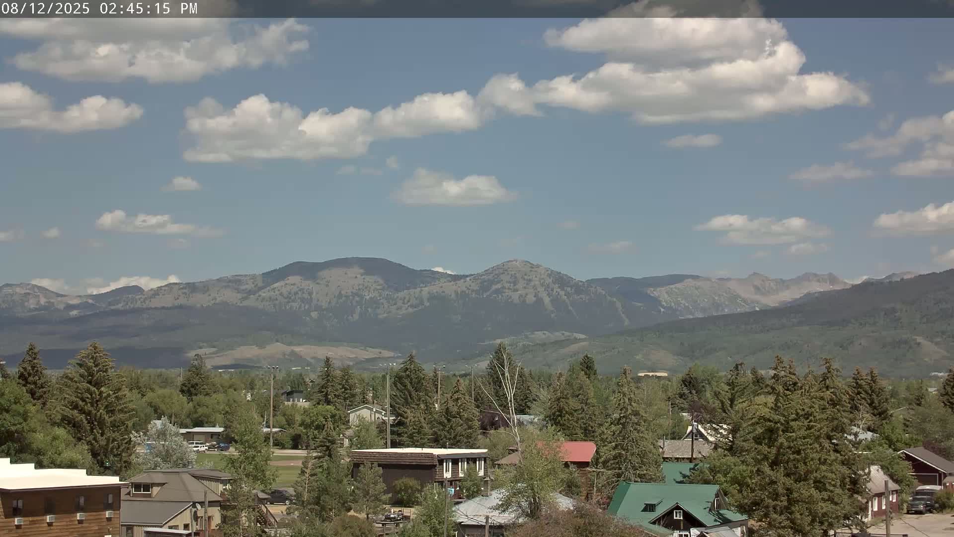 A partly cloudy sky overlooks a small town nestled among green trees, with a mountain range in the background.