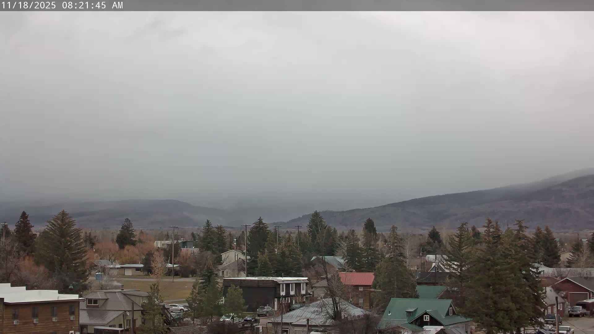 A partly cloudy sky overlooks a small town nestled among green trees, with a mountain range in the background.