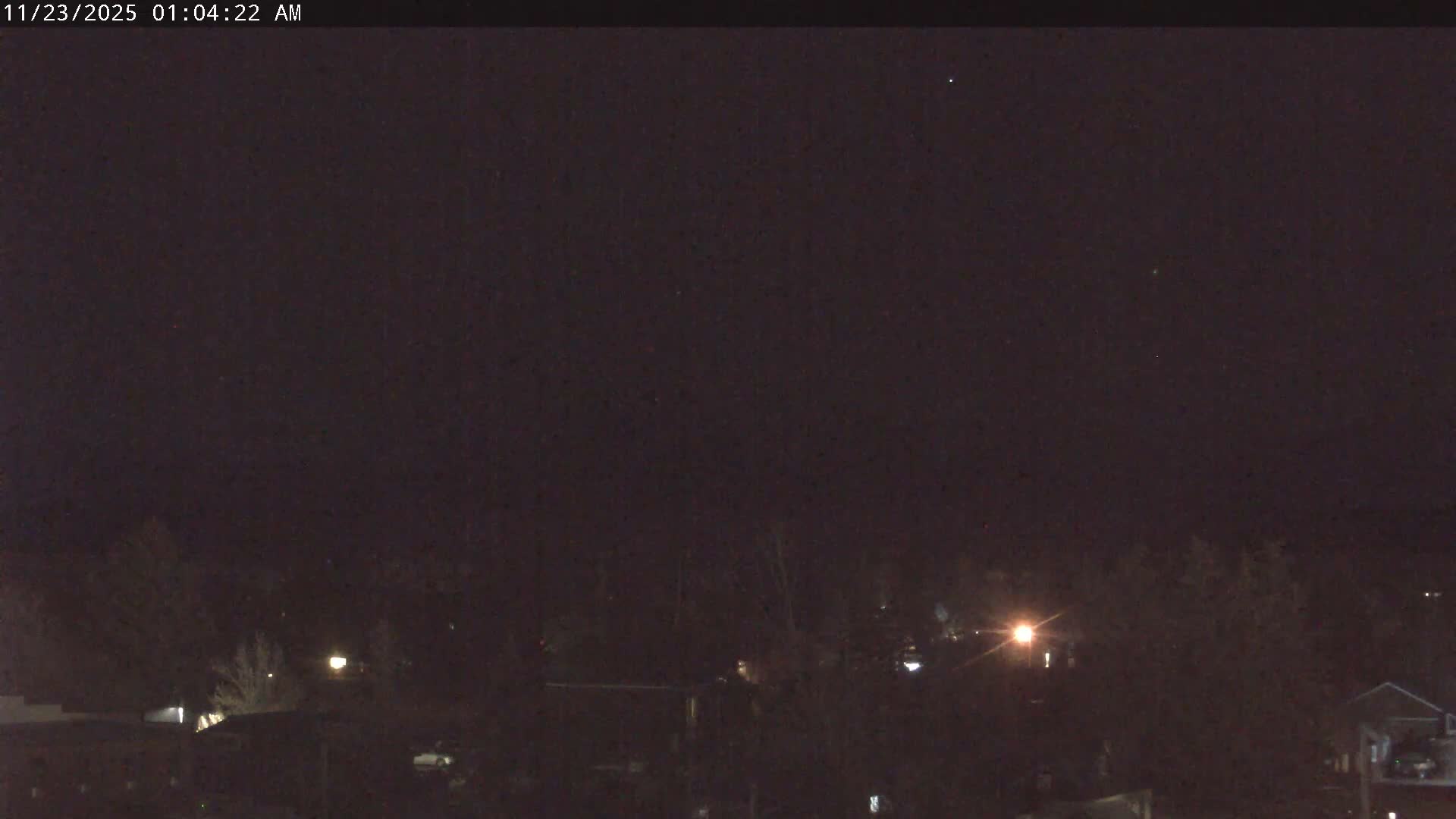 A clear night scene reveals a dark sky with a few faint stars, overlooking a distant urban or suburban landscape with scattered trees and buildings dimly lit by artificial lights.