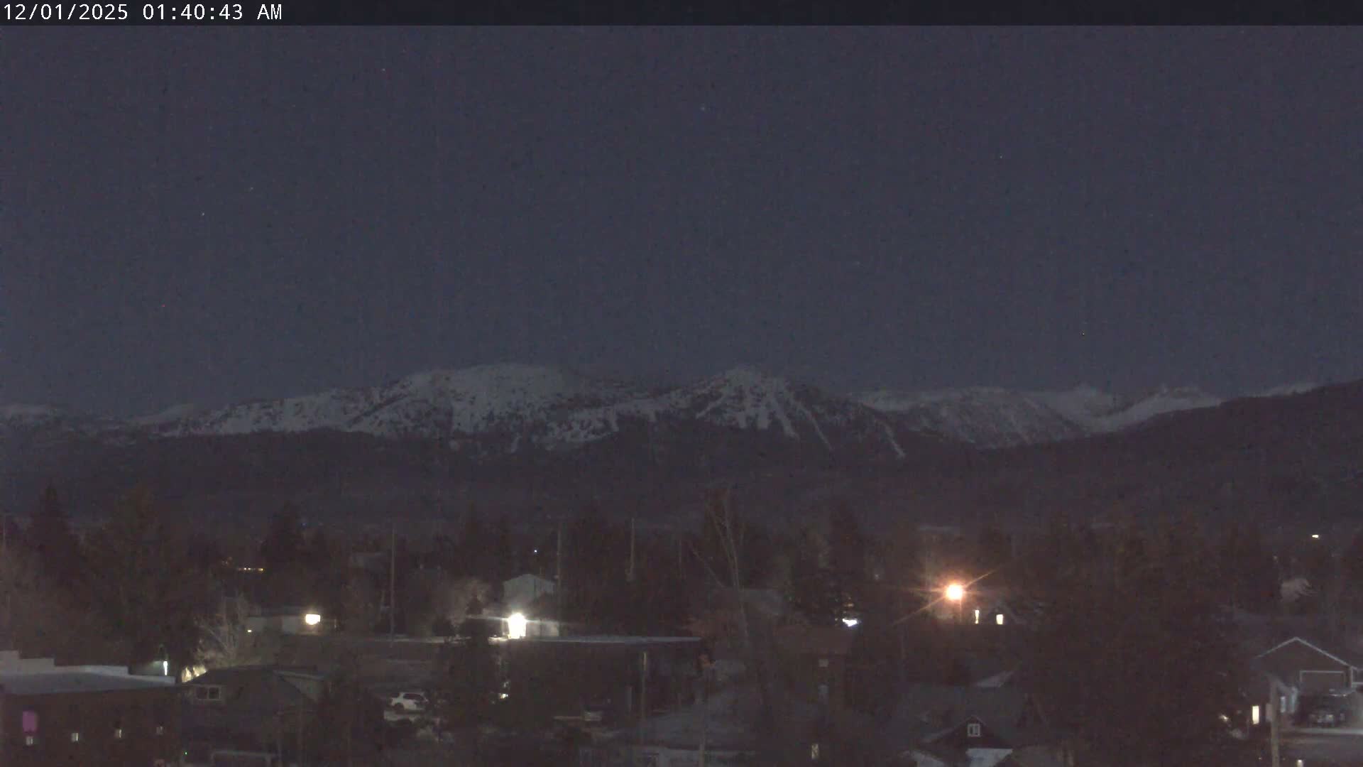A clear and cold night scene captures a town with illuminated buildings and dark trees in the foreground, backed by snow-capped mountains under a dark sky with faint stars.