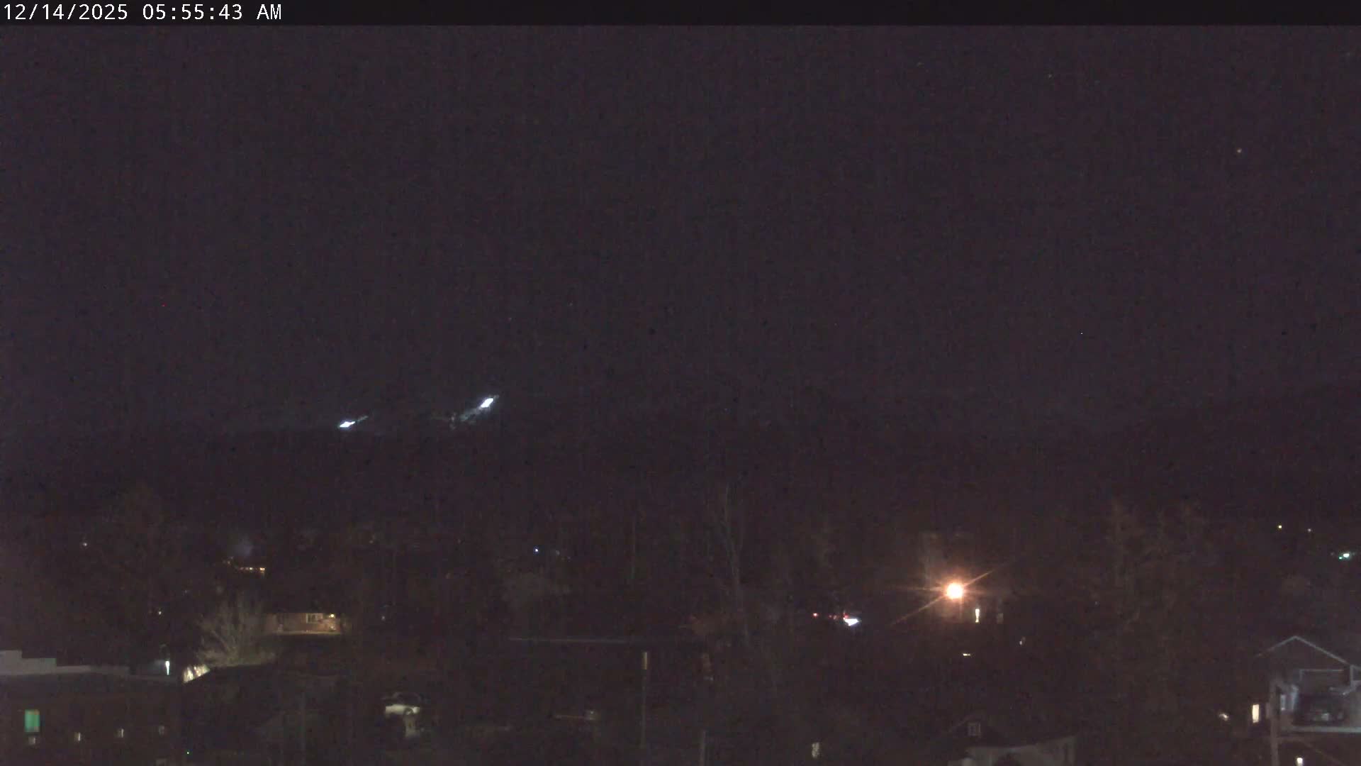 A hazy, snowy night scene shows a residential area with snow-covered houses and numerous pine trees under a dark sky, with a few distant lights visible.