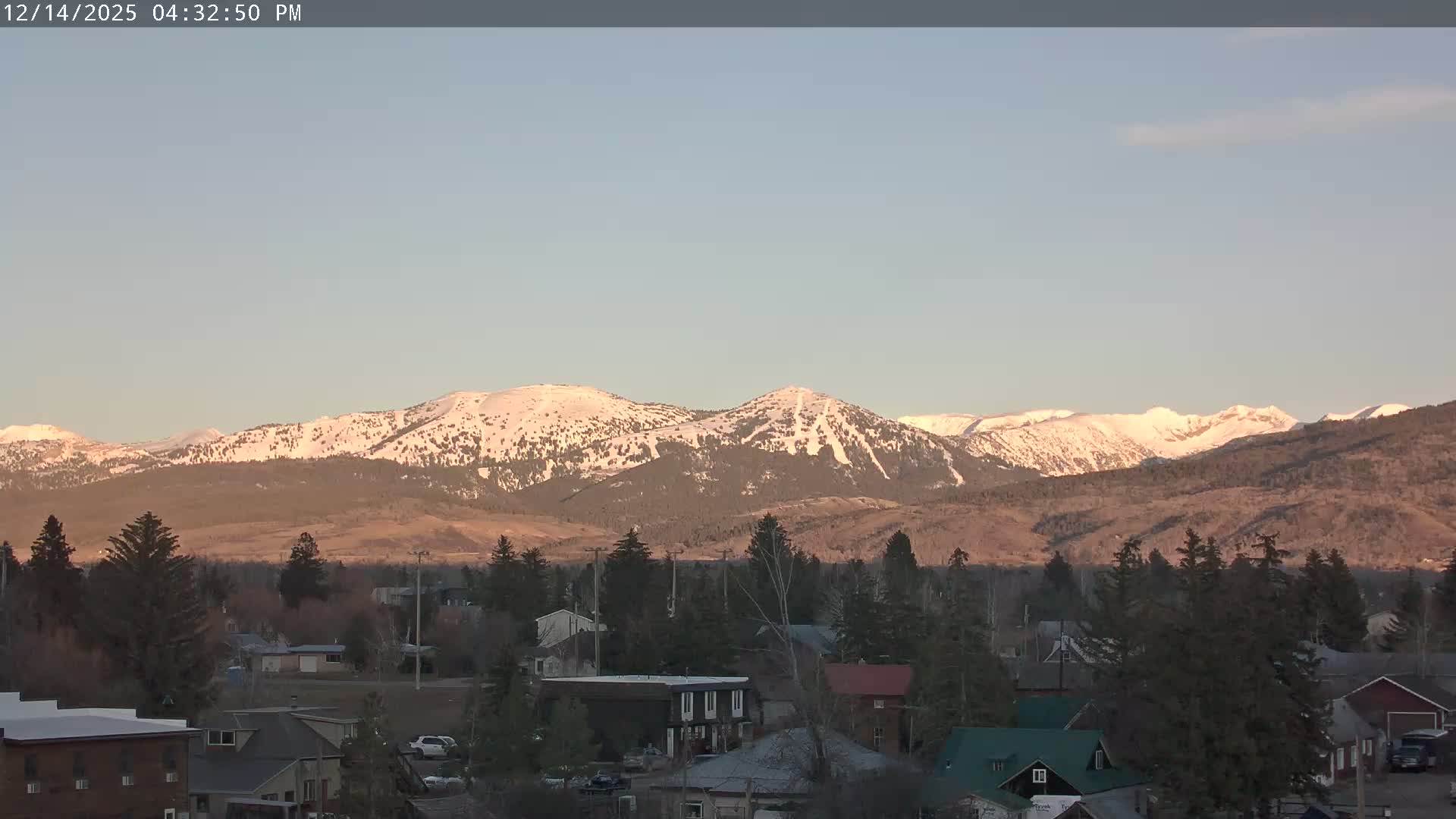A hazy, snowy night scene shows a residential area with snow-covered houses and numerous pine trees under a dark sky, with a few distant lights visible.