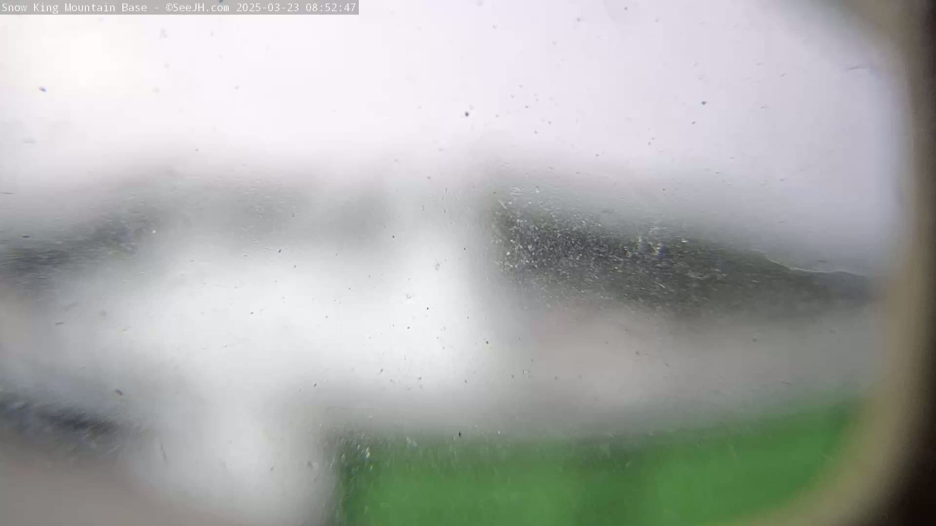 A blurry, snow-filled view of a green landscape is seen through a dirty or fogged-up window or surface.