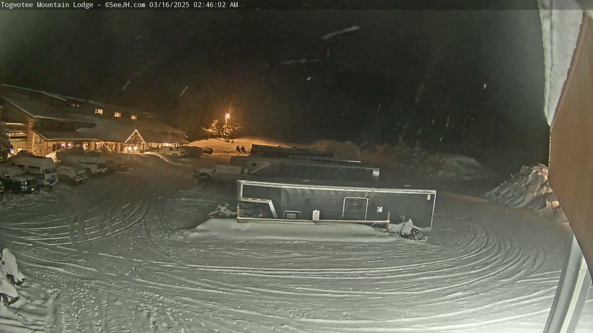 A snow-covered parking lot at night with a lodge in the background, light snowfall is occurring.