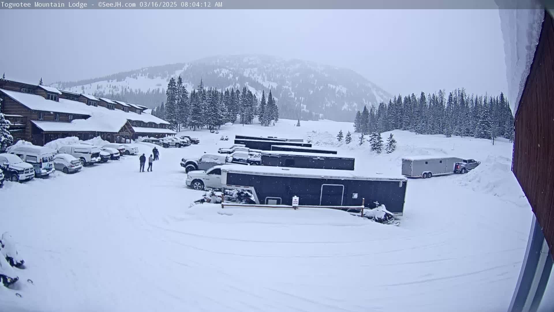 A snow-covered parking lot with vehicles and trailers in front of a snow-covered lodge, set against a backdrop of snow-covered mountains under an overcast sky.