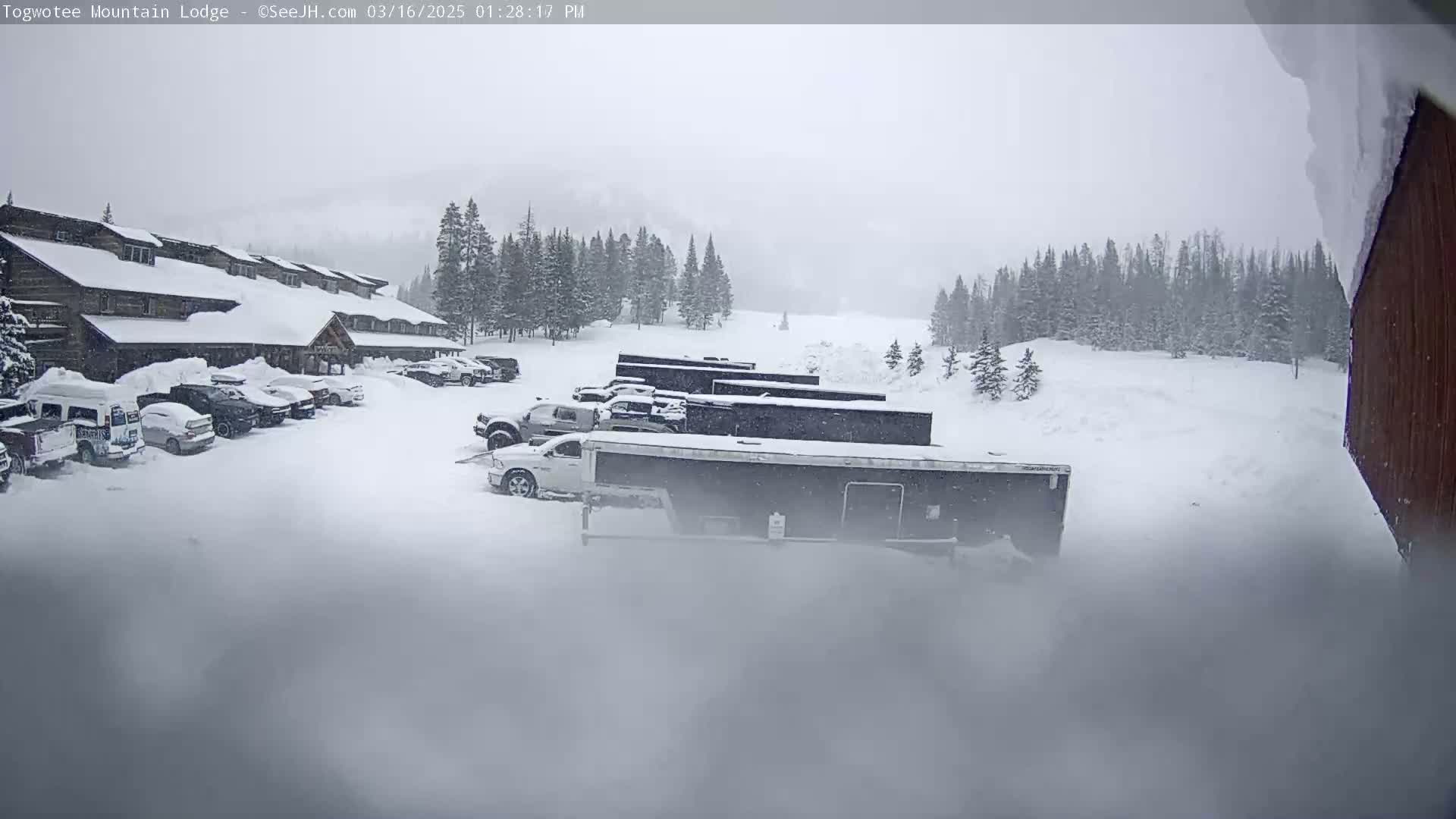 A snow-covered lodge with a parking lot full of vehicles is visible in a heavy snowstorm, with a snow-covered forest in the background.