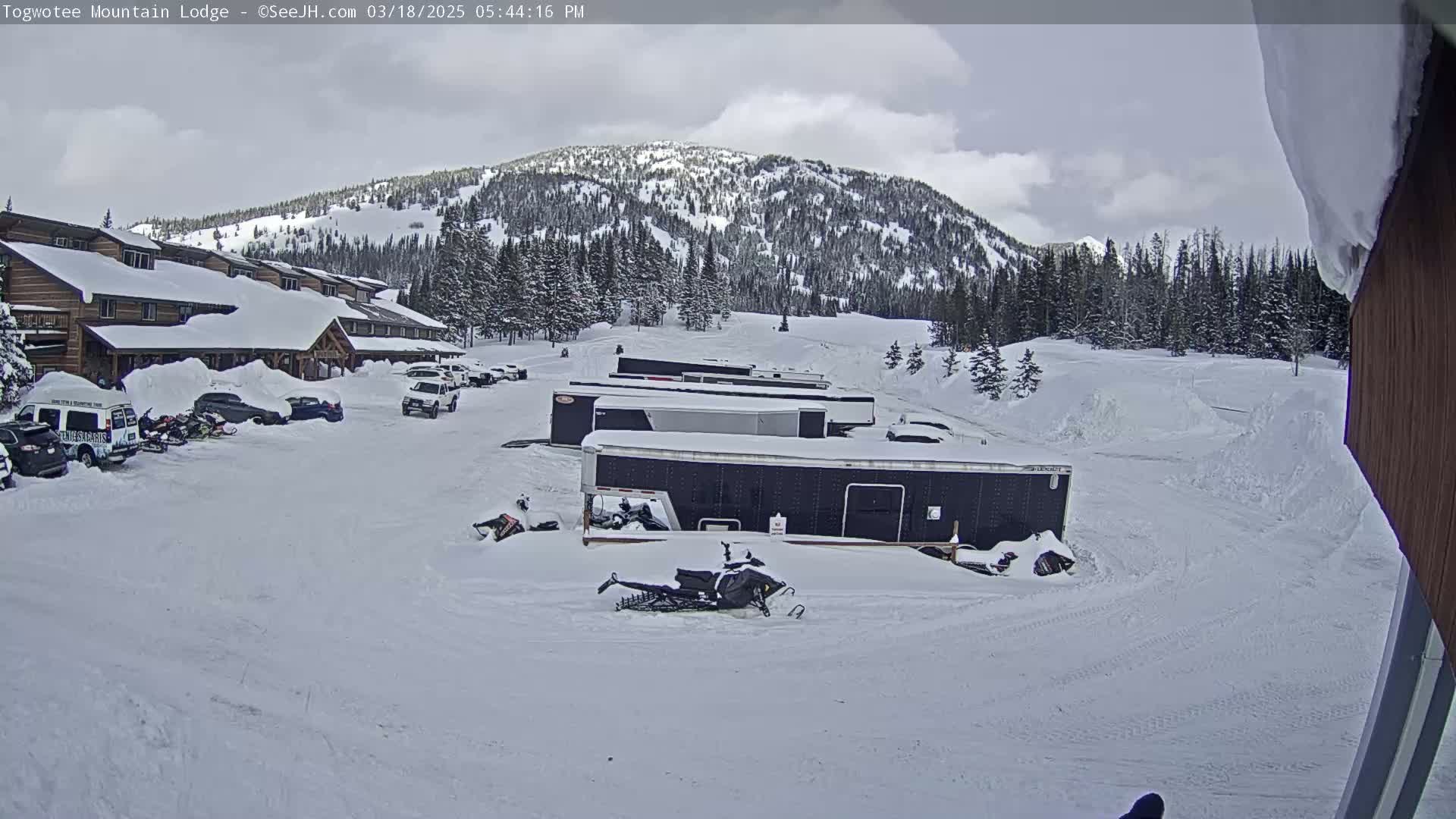 A snow-covered parking lot with several vehicles, snowmobiles, and trailers is situated in front of a snow-covered lodge, with a snow-covered mountain in the background under a partly cloudy sky.