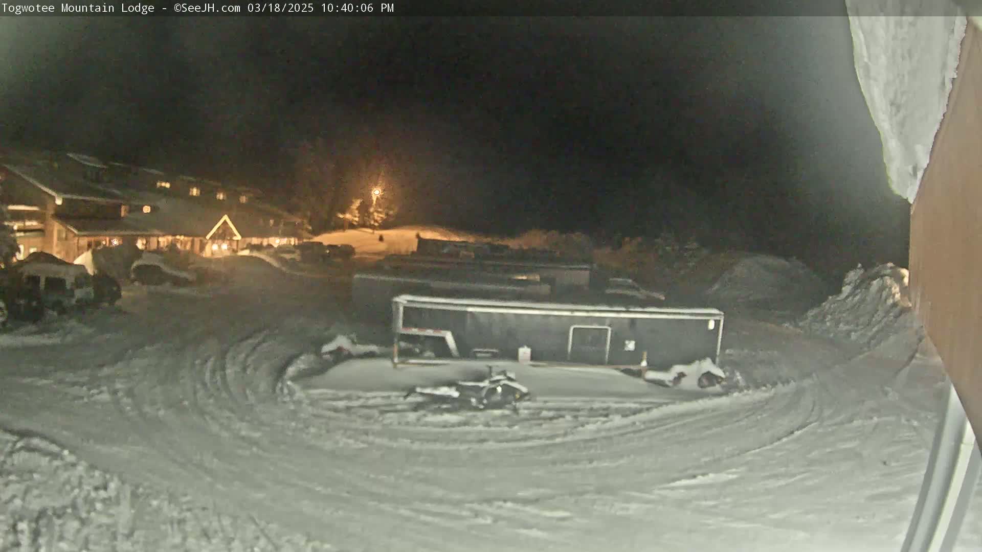 A snow-covered parking lot at night shows a lit building in the background, several vehicles, and a snowmobile parked near a large dark trailer.