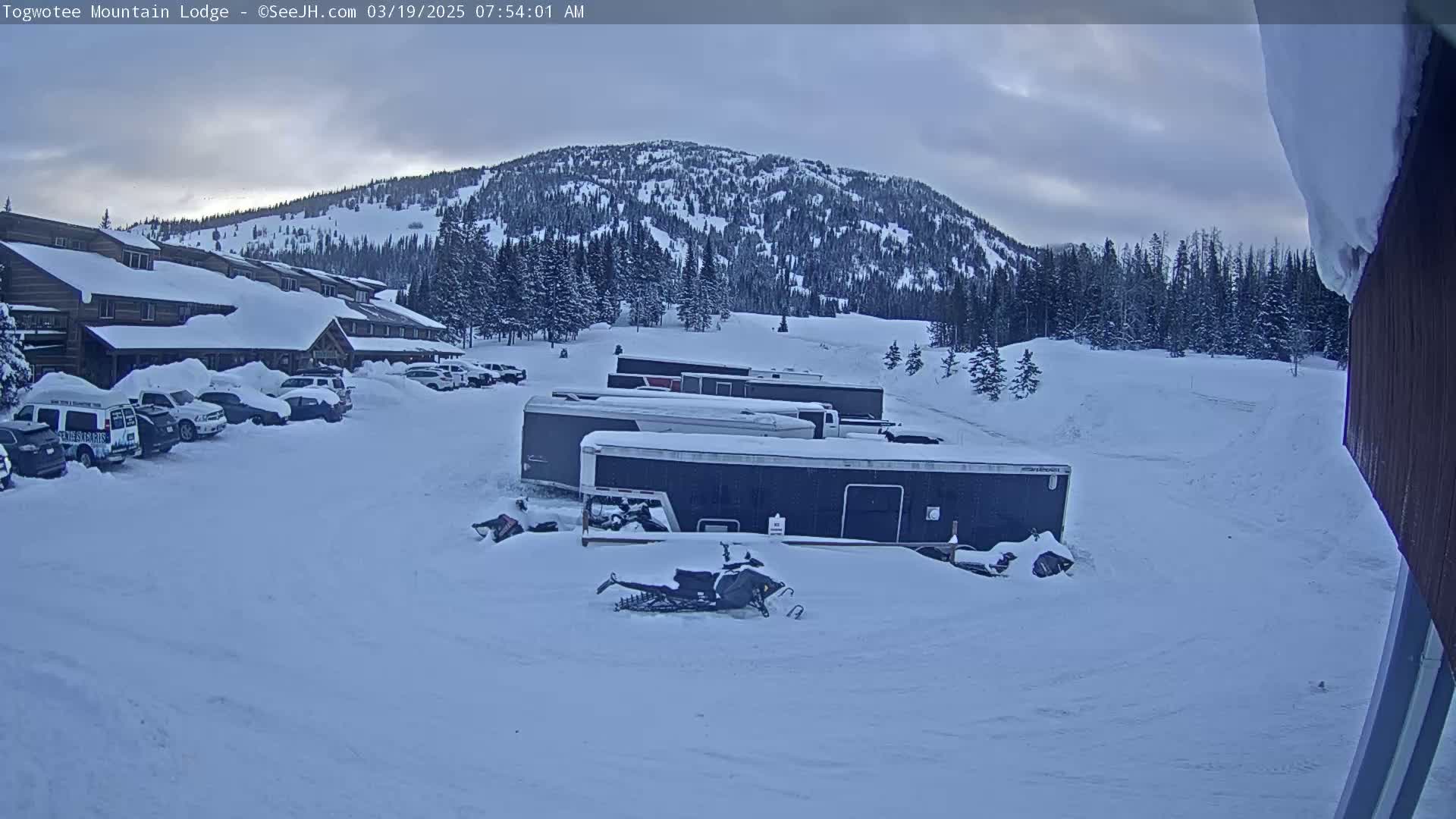 A snow-covered mountainside is visible behind a lodge and parking lot with several vehicles and snowmobiles parked in the snow, under an overcast sky.