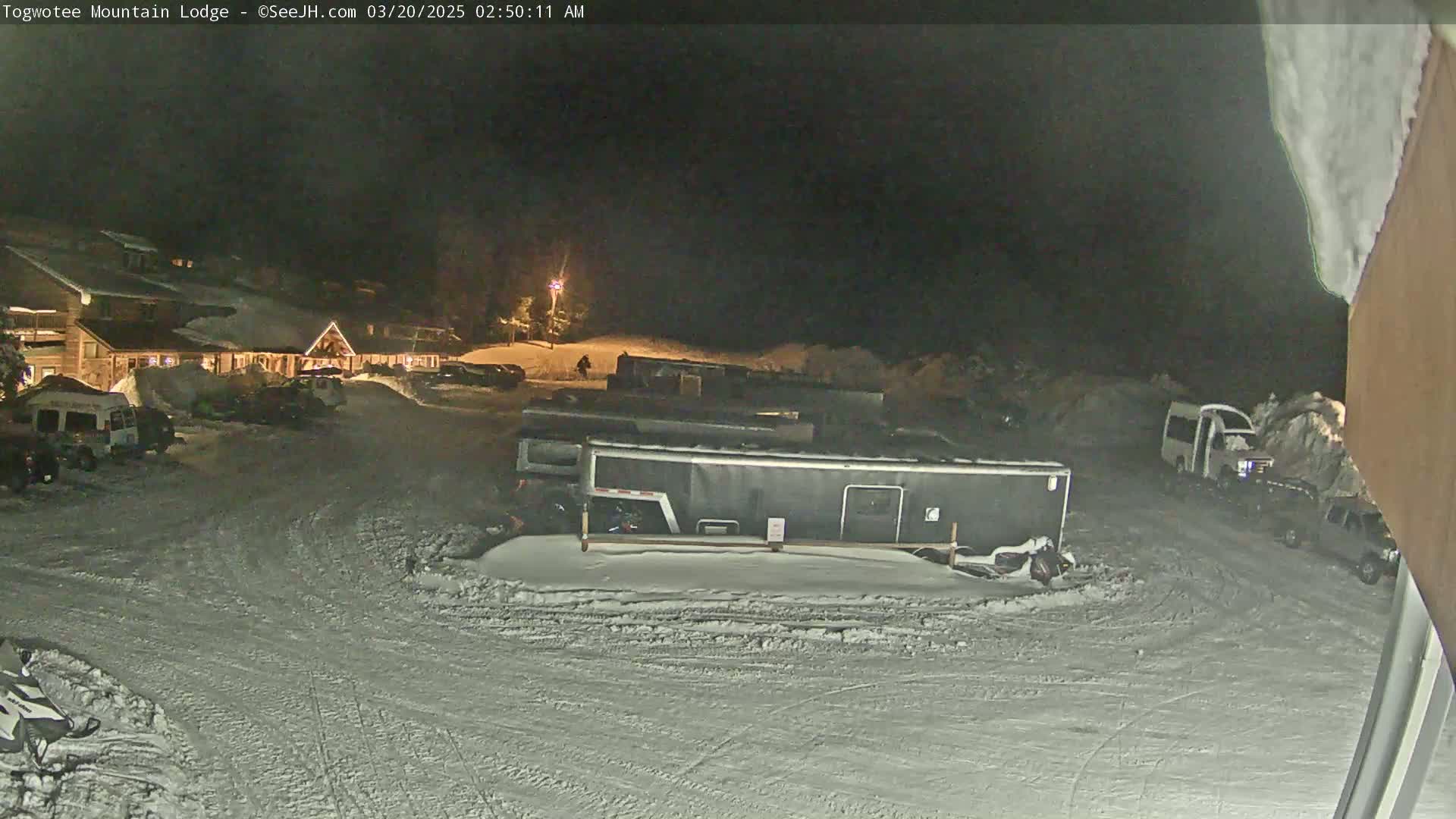 A snow-covered parking lot at night, with a lodge and several vehicles, including trailers, visible under dim lighting.