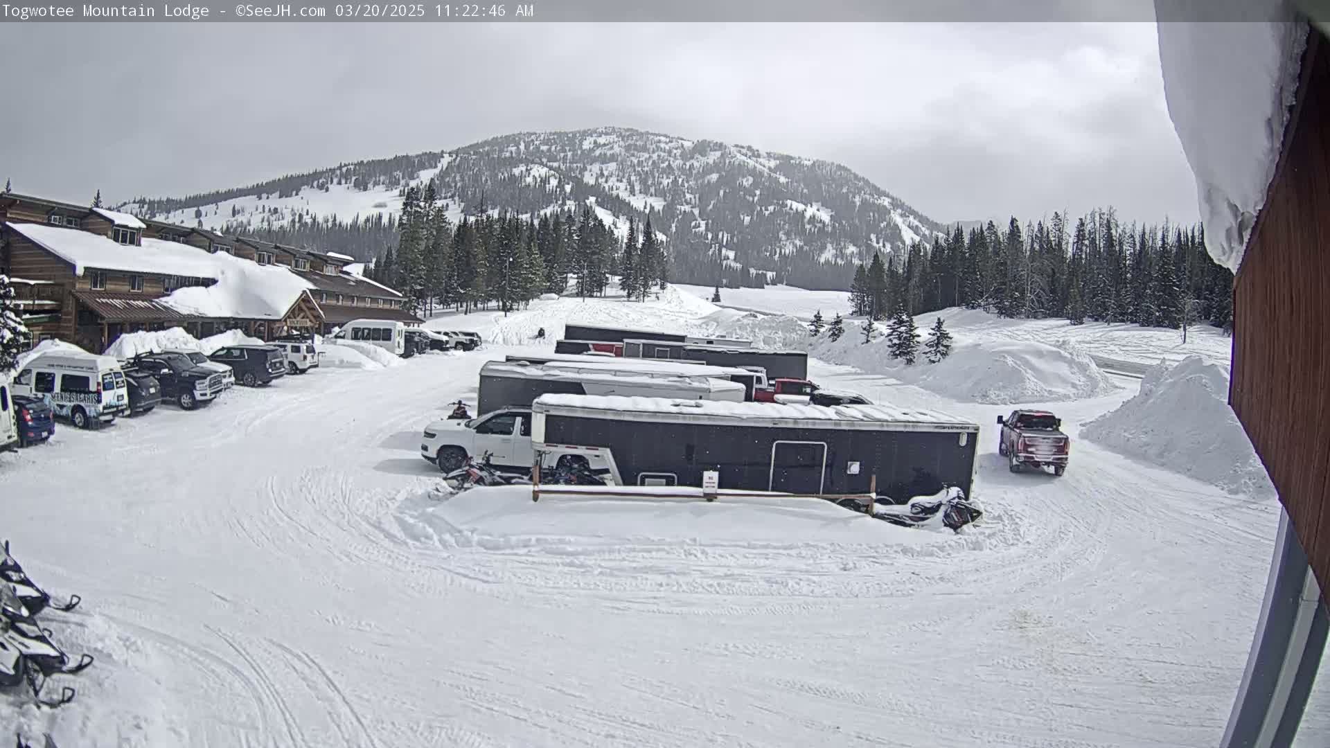 A snow-covered parking lot with several vehicles and trailers in front of a lodge, situated below a snow-dusted mountain range under an overcast sky.