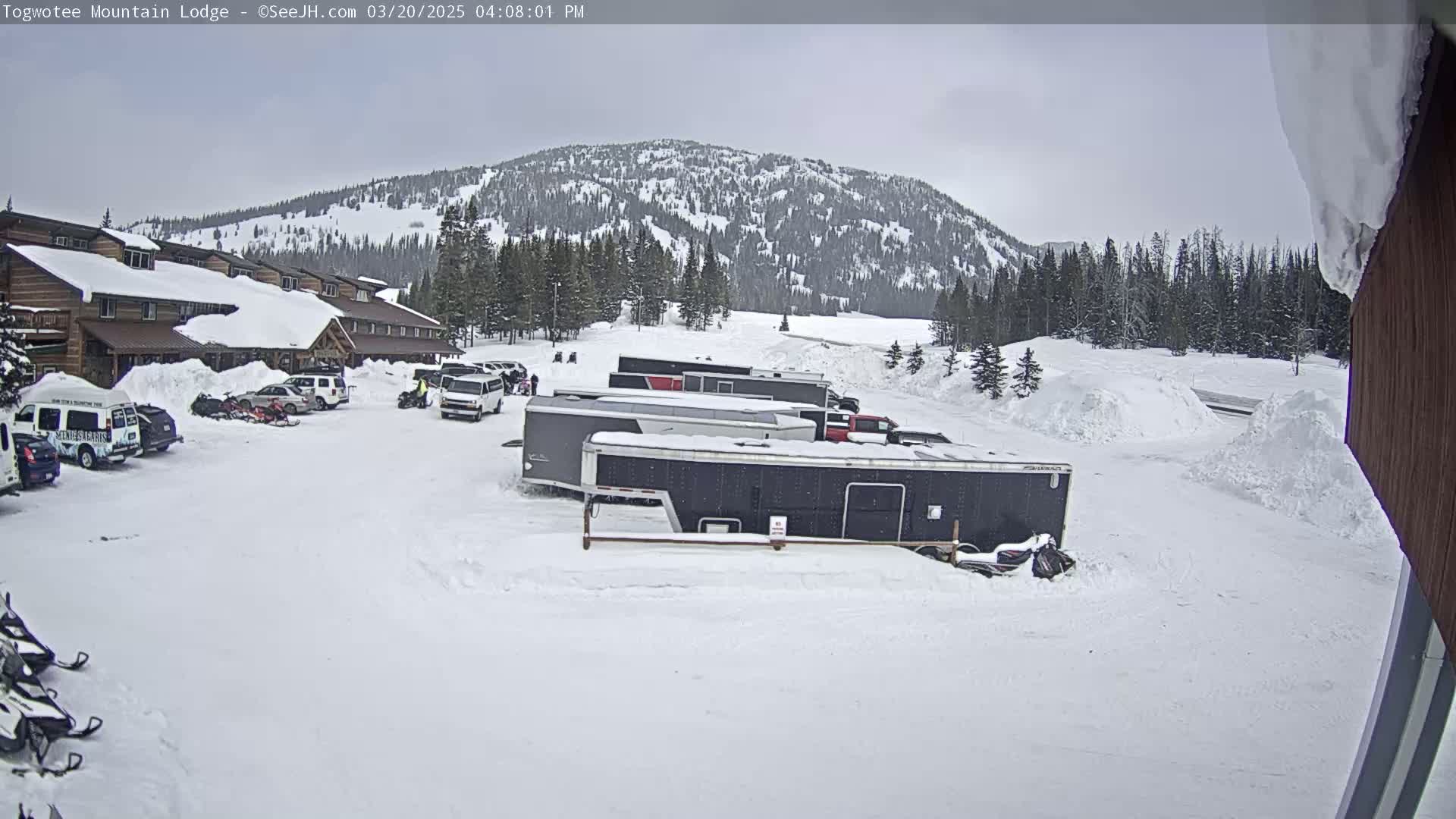 A snow-covered lodge and parking lot with several vehicles and snowmobiles are situated in front of a snow-dusted mountain range under an overcast sky.