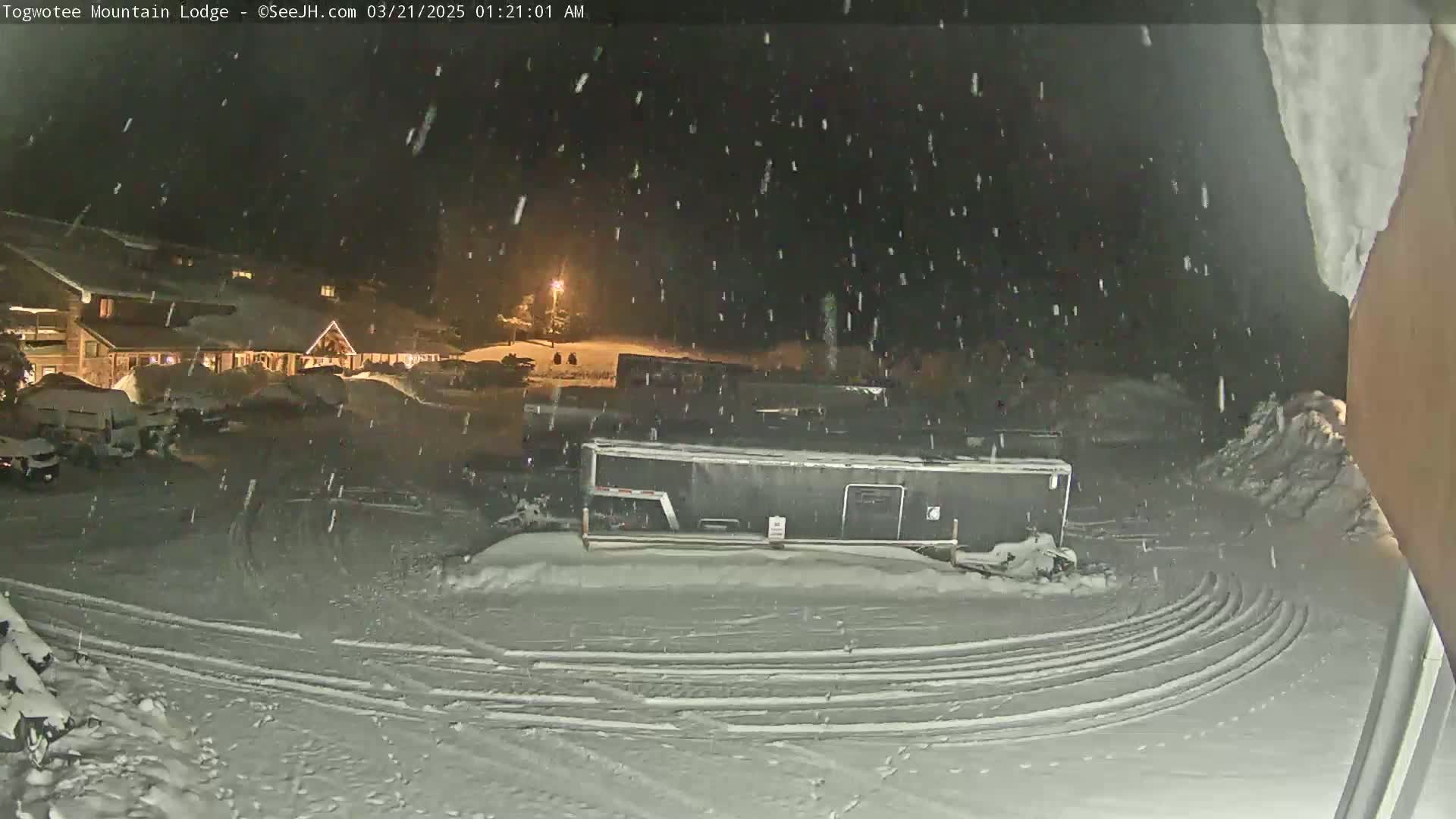 A snow-covered parking lot with a large dark trailer and a building in the background under heavy snowfall at night.
