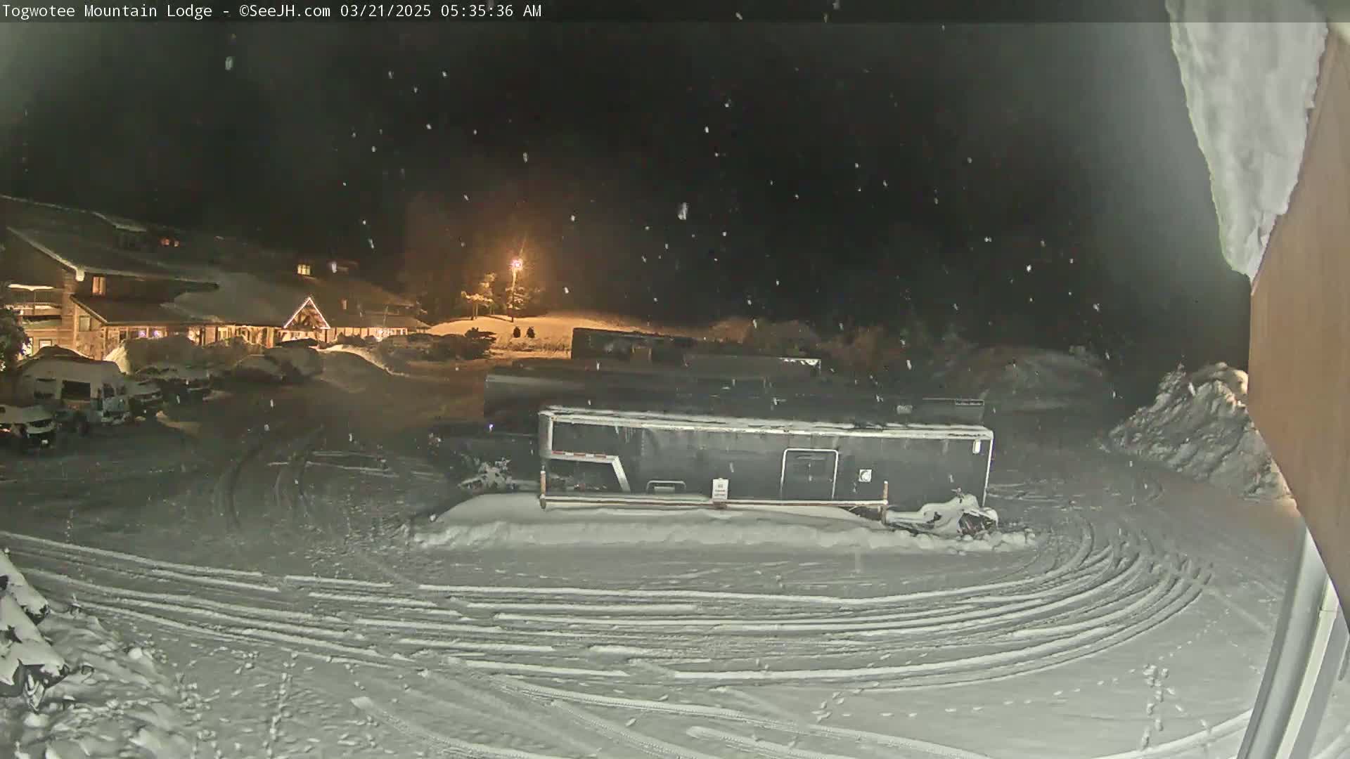 A snow-covered parking lot with a lodge and trailers in the background under a lightly falling snow at night.