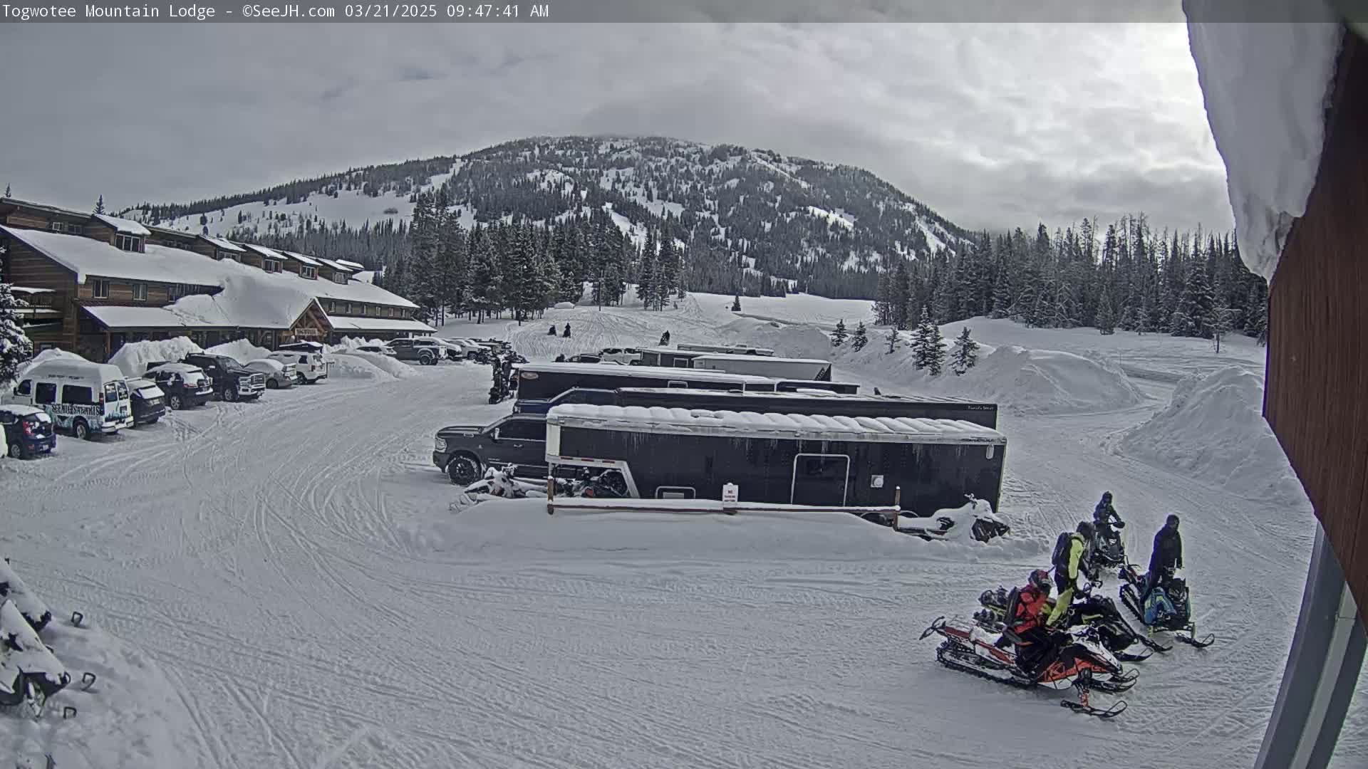 A snow-covered parking lot with several vehicles and snowmobiles, in front of a lodge and a snow-covered mountain under an overcast sky.