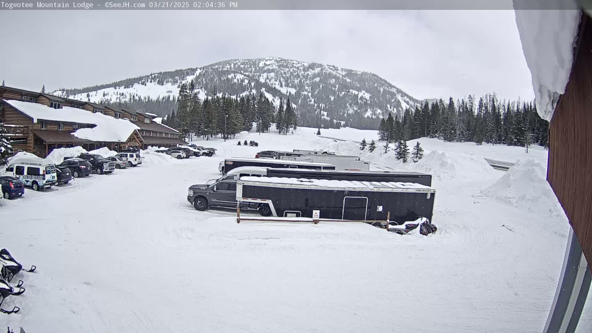 A snow-covered parking lot with several vehicles, including trucks and trailers, is situated in front of a lodge, with a snow-covered mountain in the background under an overcast sky.