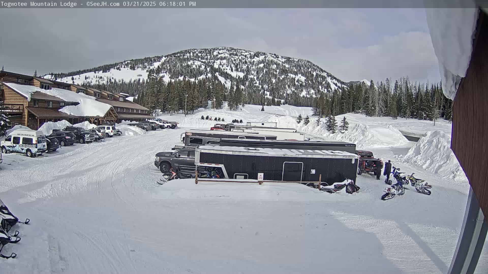 A snow-covered parking lot with cars, snowmobiles, and trailers in front of a lodge, situated below a snow-dusted mountain under a partly cloudy sky.