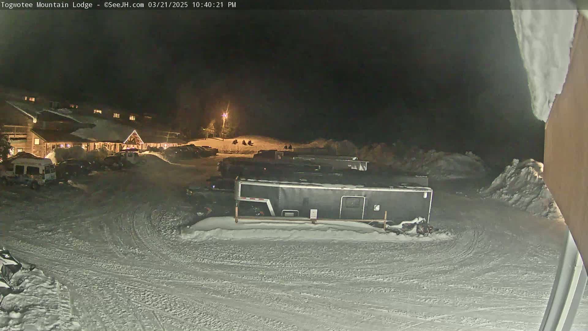 A snow-covered parking lot at night, illuminated by artificial lights, with a lodge in the background and several vehicles parked, including a large enclosed trailer.