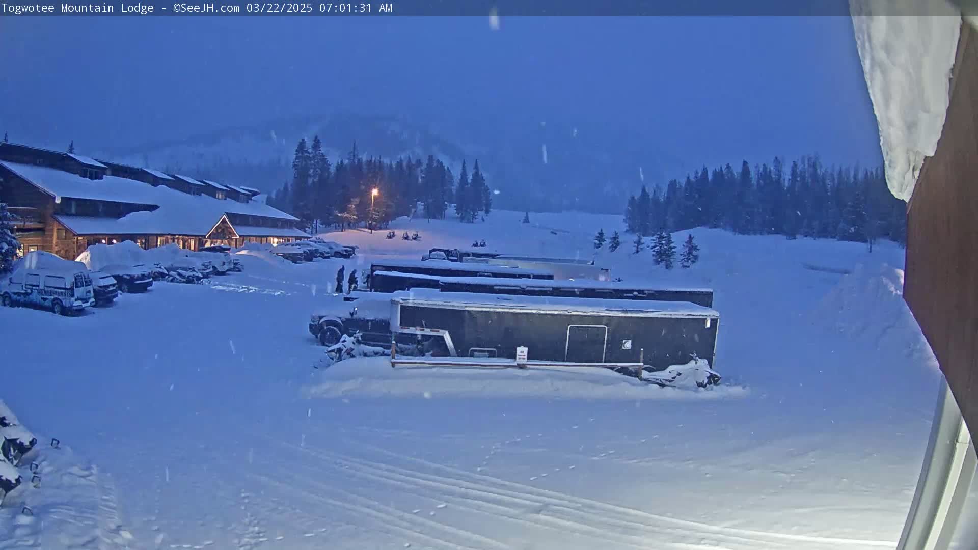 A snow-covered lodge parking lot with several vehicles and trailers under a twilight sky, with light snowfall.
