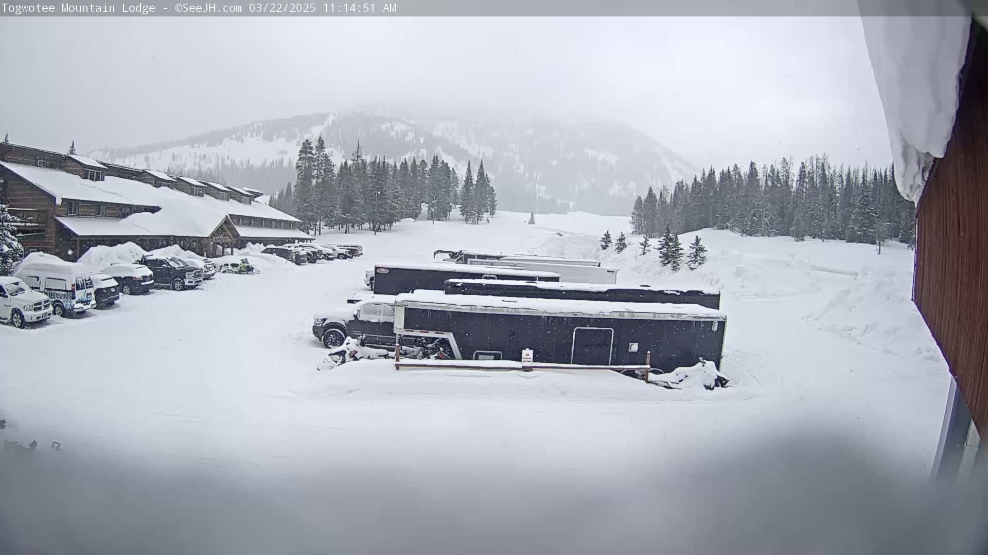 A snow-covered parking lot with several vehicles and trailers in front of a lodge, under a gray, overcast sky with snow falling on a mountain background.