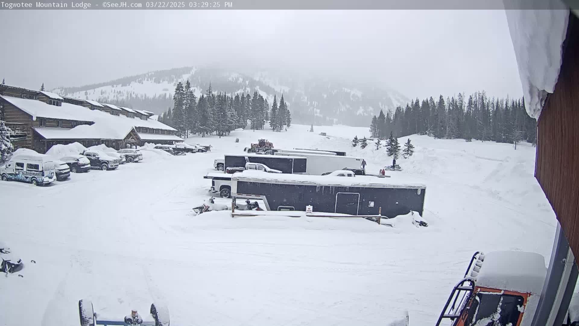 A snow-covered lodge and parking lot with several vehicles and trailers are visible in a snowy landscape, under an overcast sky.