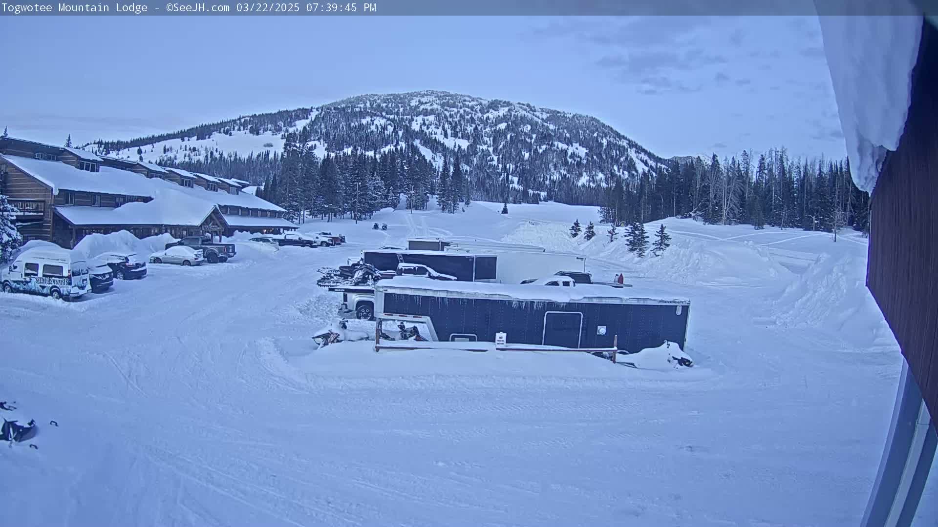 A snow-covered parking lot with several vehicles and trailers sits in front of a multi-story lodge, with a snow-covered mountain in the background under a partly cloudy sky.