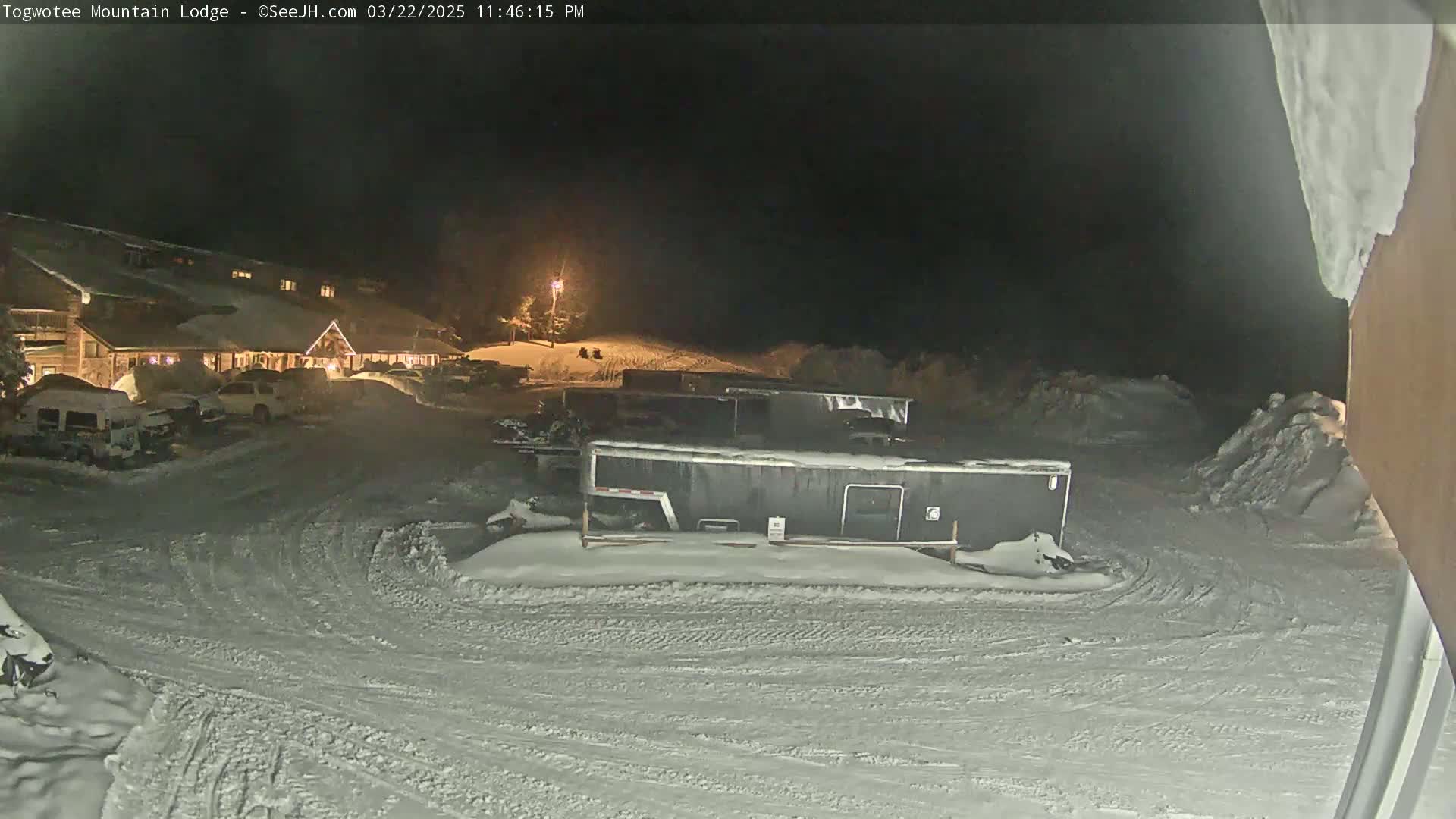 A snow-covered parking lot at night, illuminated by a single light, shows a lodge, several vehicles, and large dark containers.