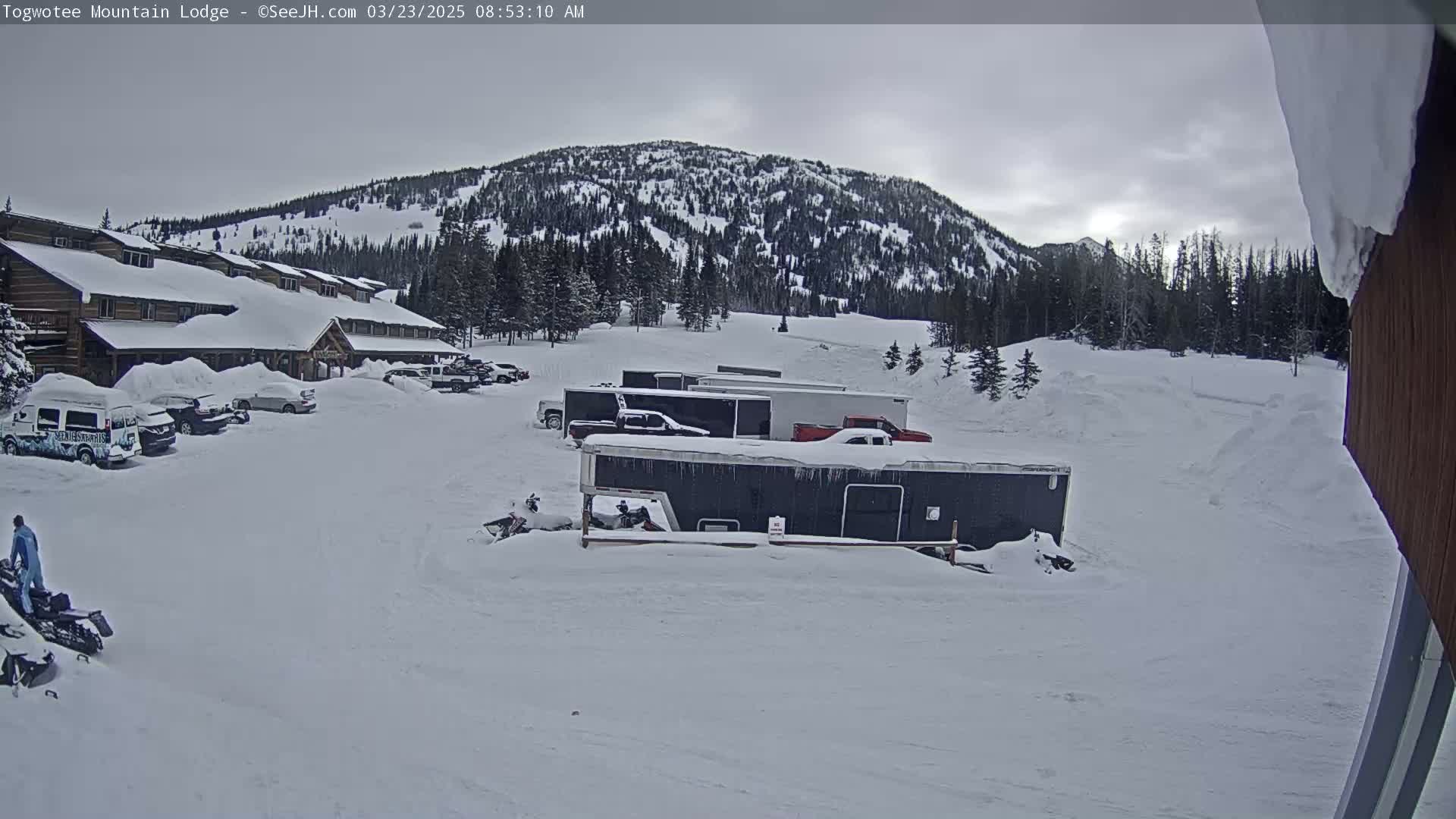 A snow-covered parking lot with several vehicles and trailers in front of a lodge, all situated below a snow-dusted mountain under an overcast sky.
